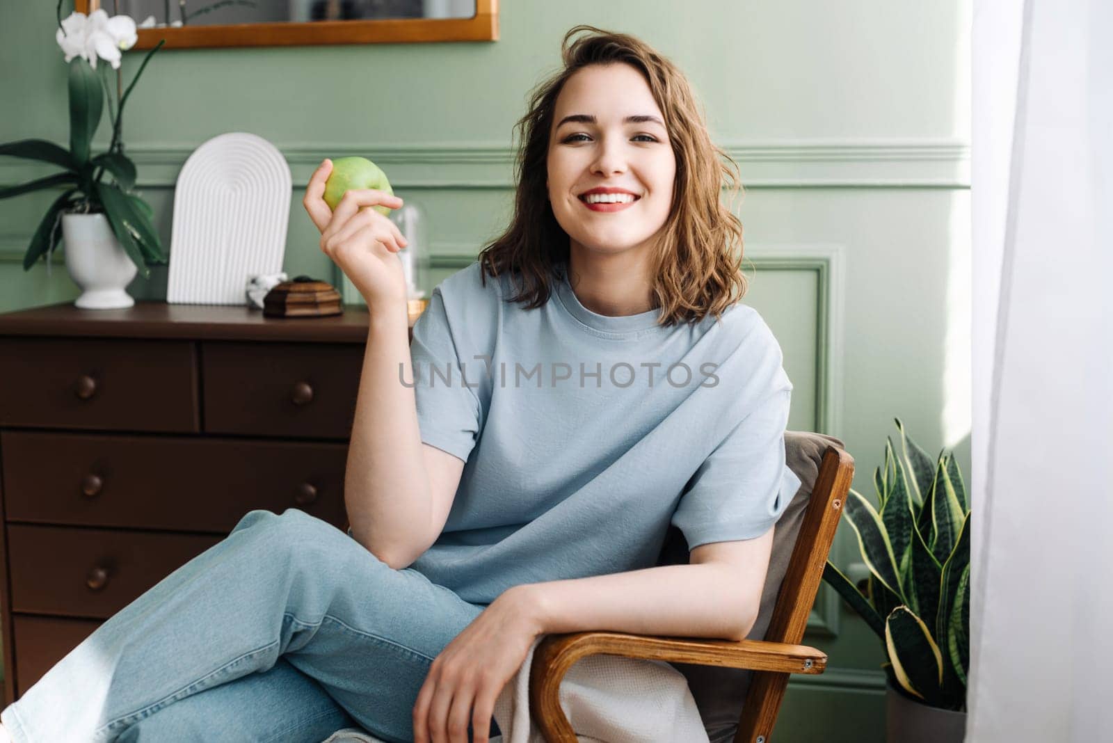 Joyful Young Woman Relaxing on Couch with Apple in Living Room. Cheerful Woman Sitting on Sofa Enjoying Apple in Living Room Scene. Leisure Time at Home.