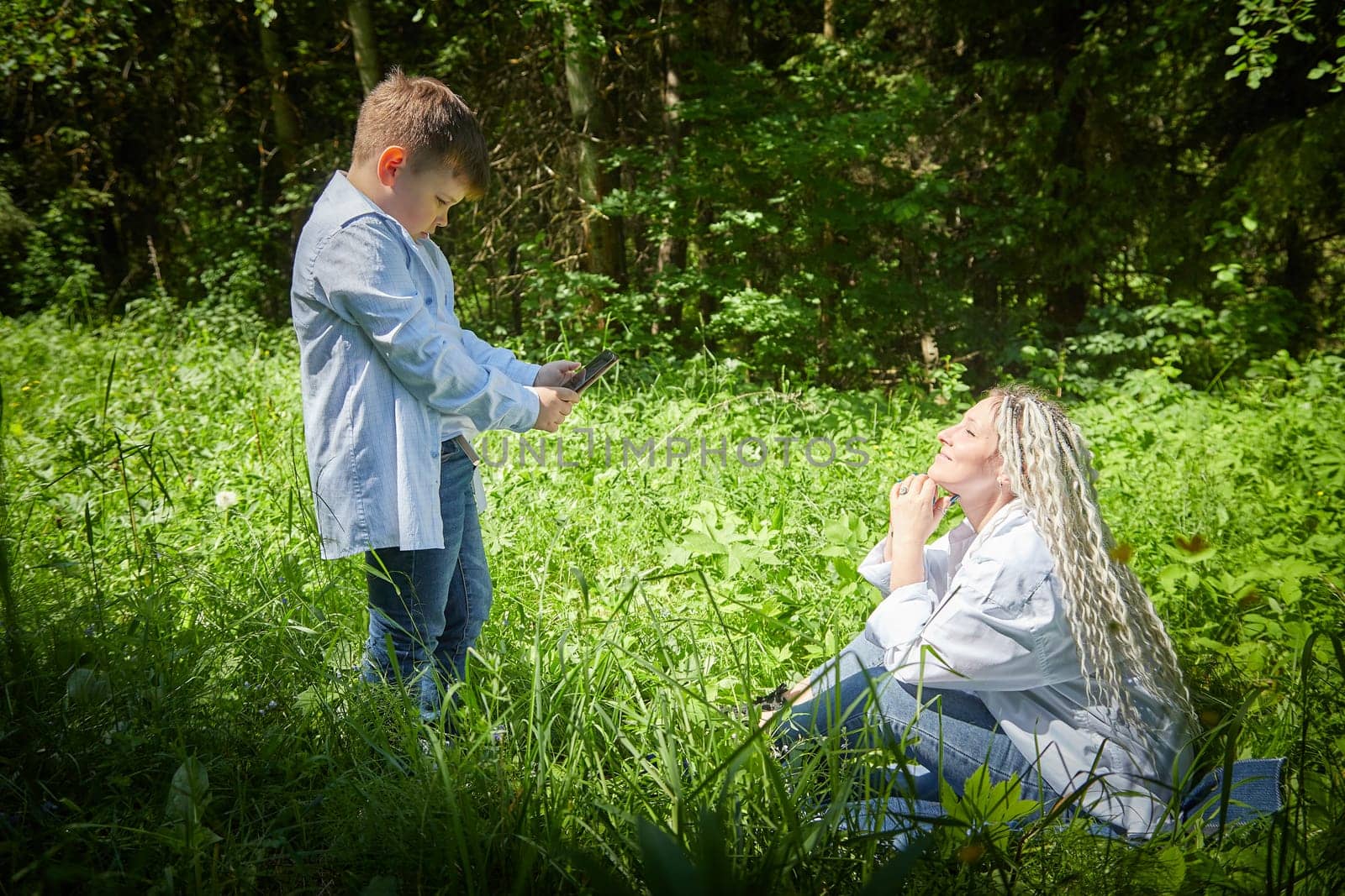A young teenage son takes pictures of his mother on a cell phone in nature. Mom and boy having fun in the grass in clearing