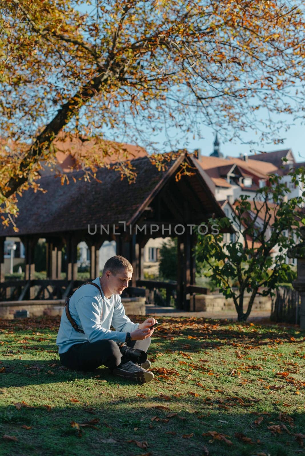 Professional Photographer Taking Picture Of Beautiful Autumn Park. Man Professional Photographer Sit With Camera And With Smartphone In Autumn Park. Retouched, Vibrant Colors, Brownish Tones. by Andrii_Ko