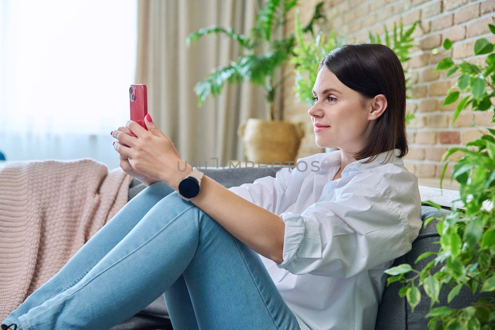 Young smiling happy woman using smartphone, sitting on sofa at home in living room. Internet online services, communication, mobile applications, technologies for leisure study work lifestyle