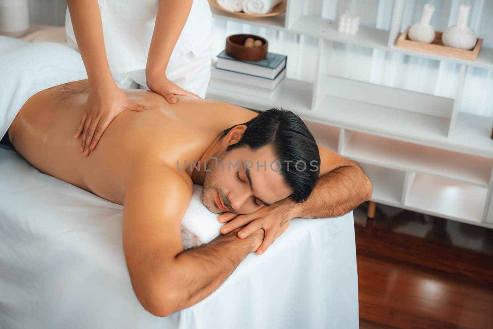 Caucasian man customer enjoying relaxing anti-stress spa massage and pampering with beauty skin recreation leisure in day light ambient salon spa at luxury resort or hotel. Quiescent