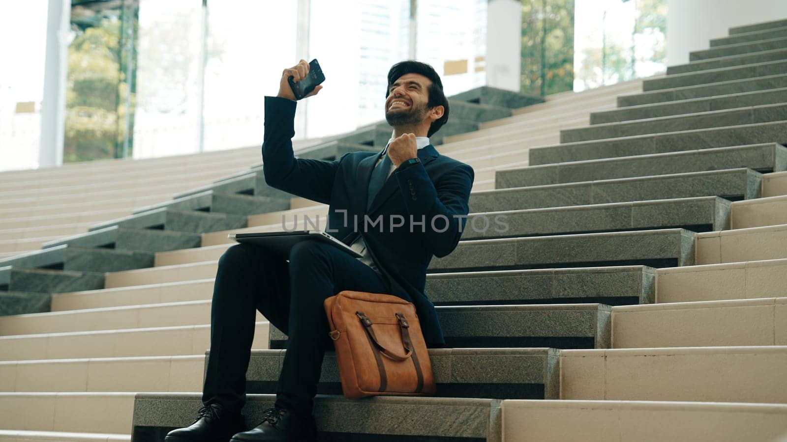 Successful business man celebrate successful project while sitting at stairs. Smart project manager getting new gob, getting promotion, increasing sales while calling friends by using phone. Exultant.