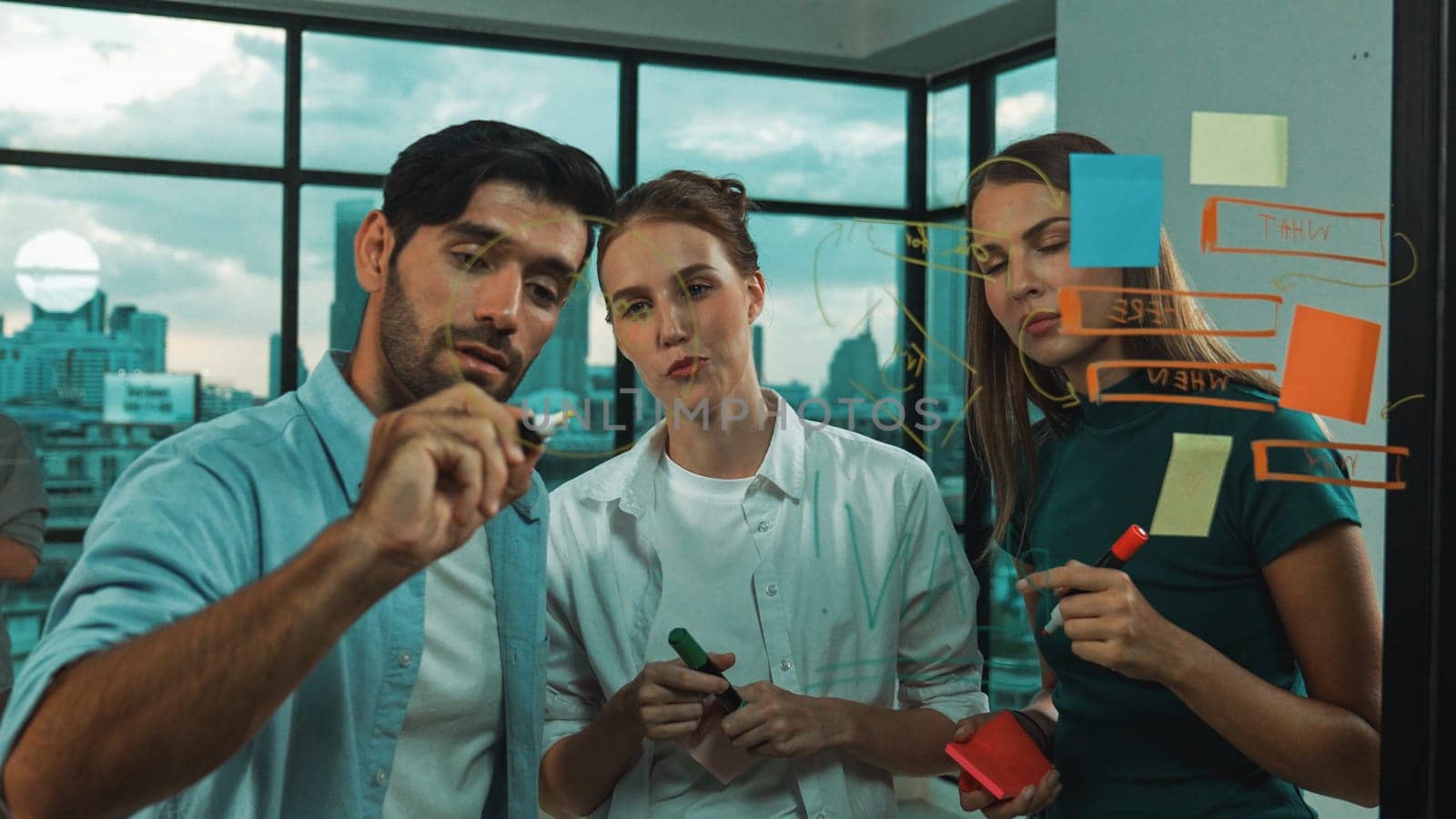 Professional business team brainstorm, sharing, thinking idea while skilled male leader write on sticky notes at glass wall. Skilled manager placed business plan on glass wall at office. Tracery