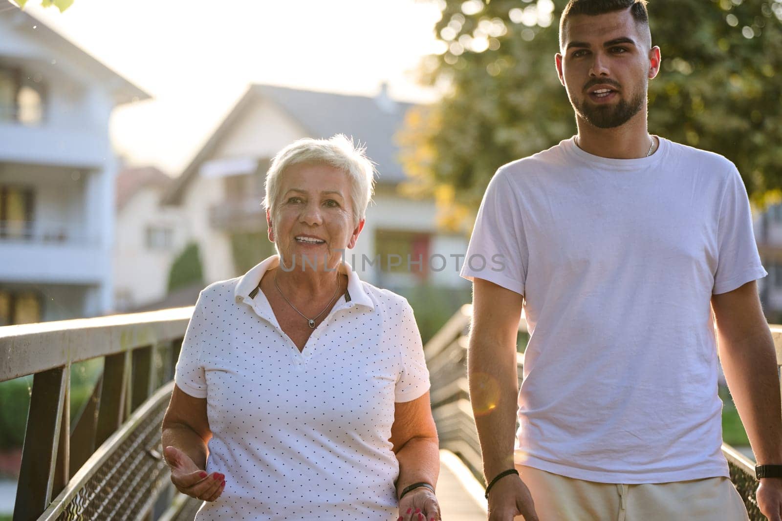 A handsome man and an older woman share a serene walk in nature, crossing a beautiful bridge against the backdrop of a stunning sunset, embodying the concept of a healthy and vibrant intergenerational life