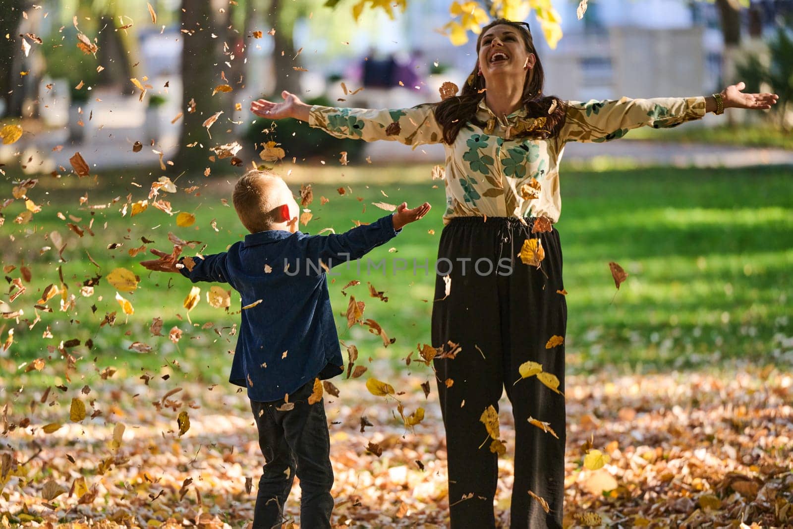 A modern woman joyfully plays with her son in the park, tossing leaves on a beautiful autumn day, capturing the essence of family life and the warmth of mother-son bonding in the midst of the fall season.