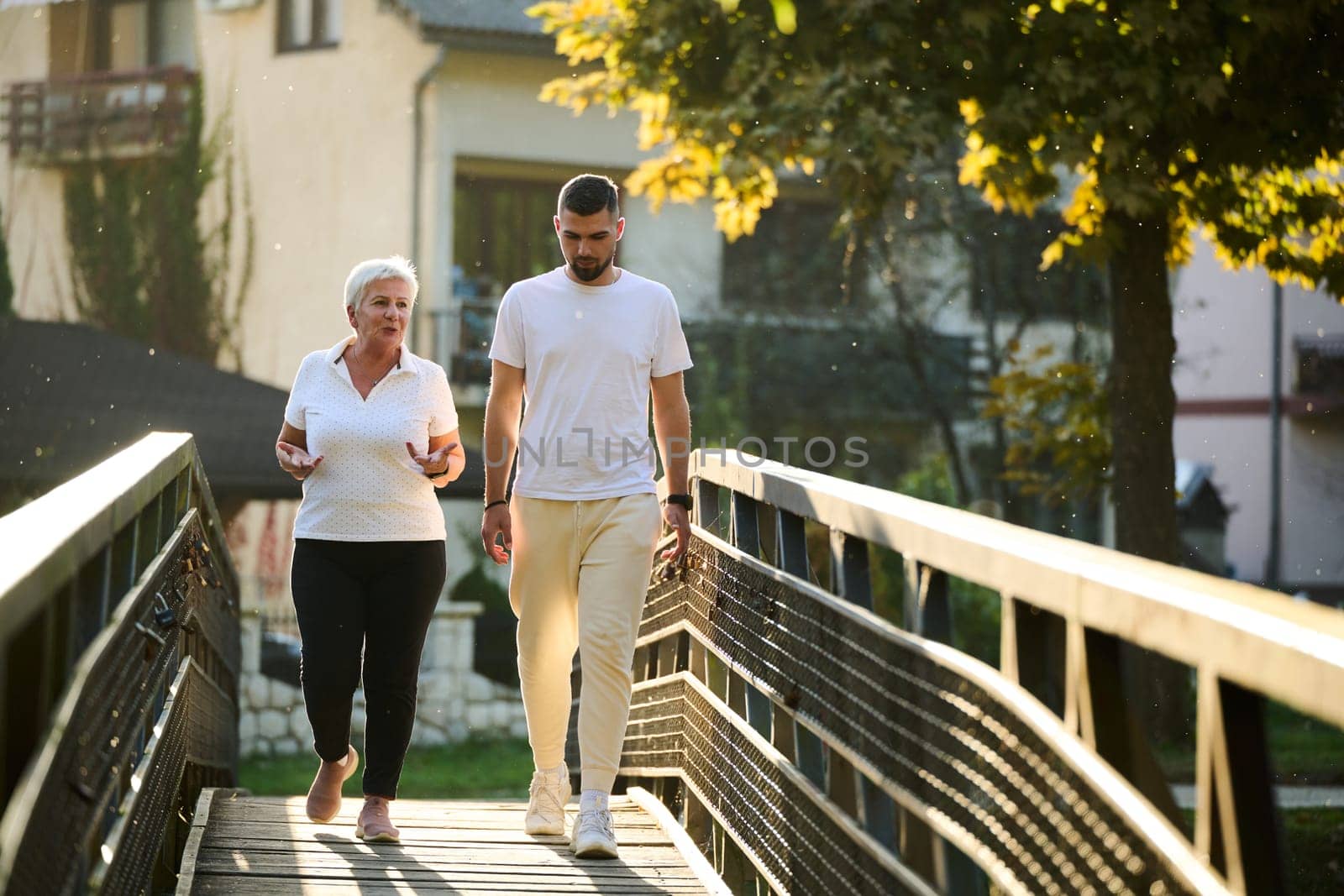 A handsome man and an older woman share a serene walk in nature, crossing a beautiful bridge against the backdrop of a stunning sunset, embodying the concept of a healthy and vibrant intergenerational life