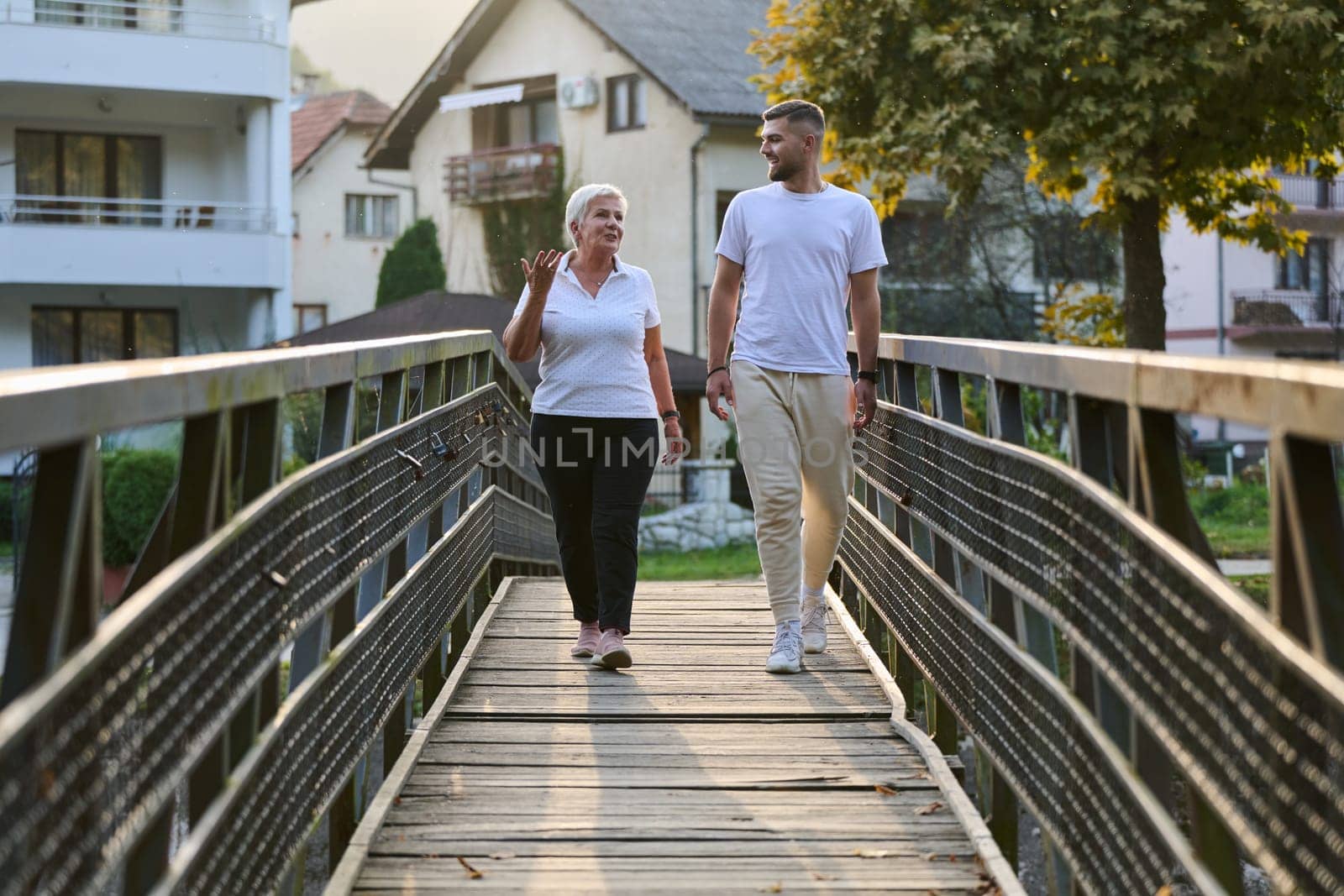 A handsome man and an older woman share a serene walk in nature, crossing a beautiful bridge against the backdrop of a stunning sunset, embodying the concept of a healthy and vibrant intergenerational life