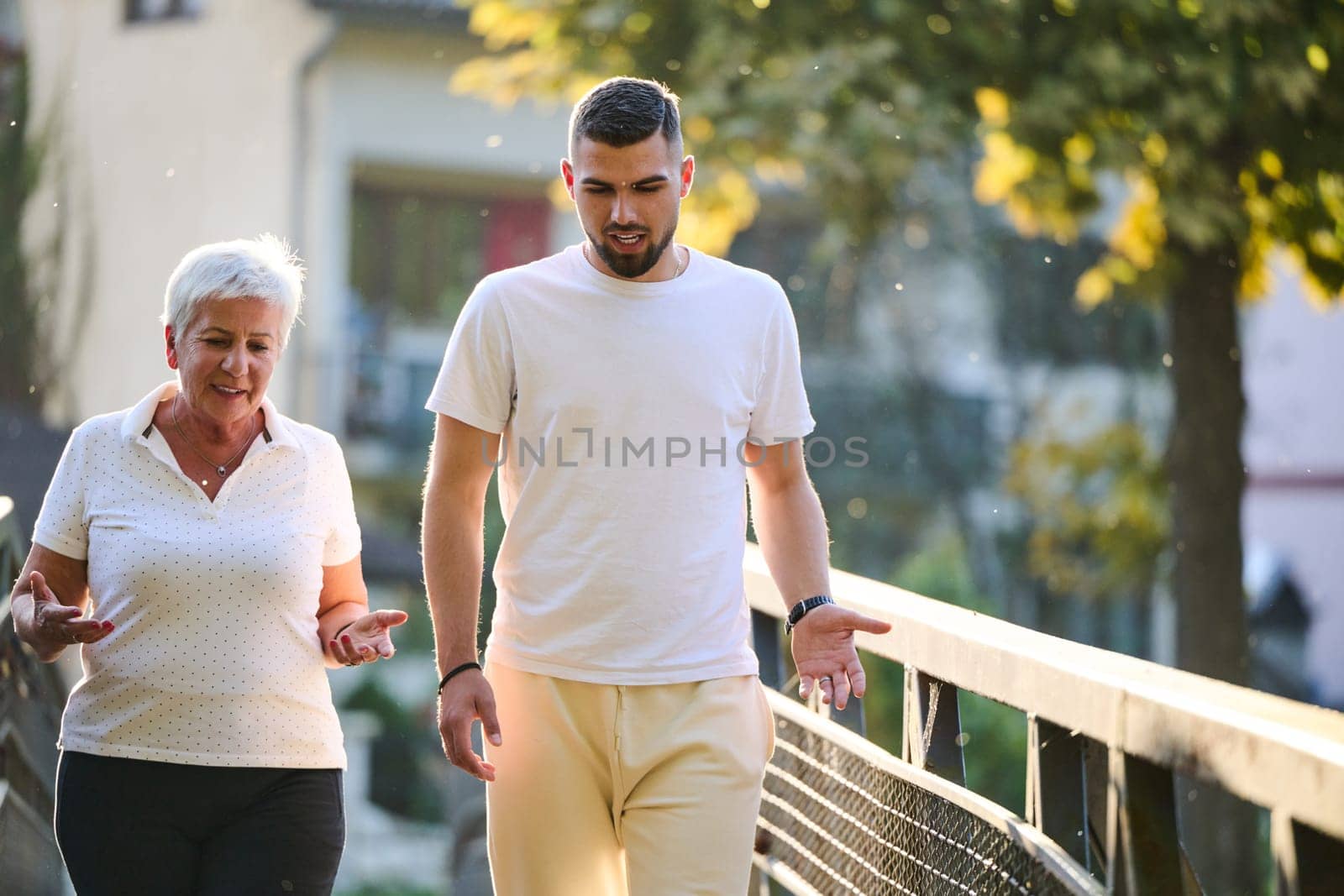 A handsome man and an older woman share a serene walk in nature, crossing a beautiful bridge against the backdrop of a stunning sunset, embodying the concept of a healthy and vibrant intergenerational life