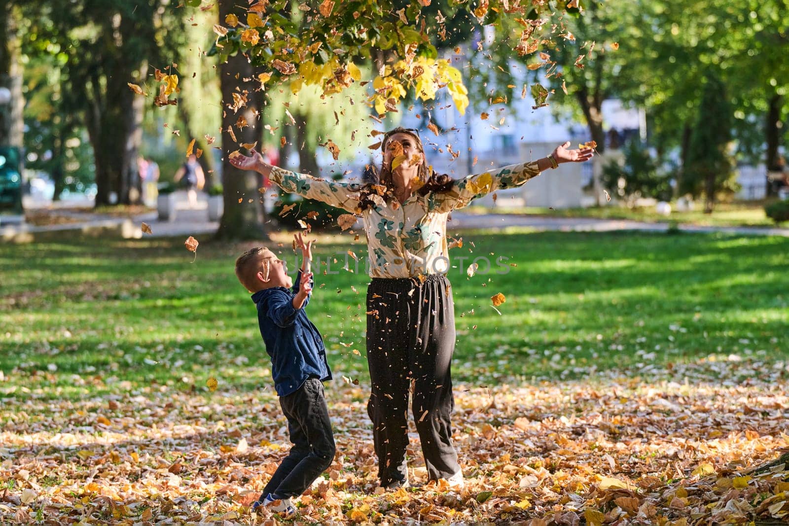 A modern woman joyfully plays with her son in the park, tossing leaves on a beautiful autumn day, capturing the essence of family life and the warmth of mother-son bonding in the midst of the fall season by dotshock