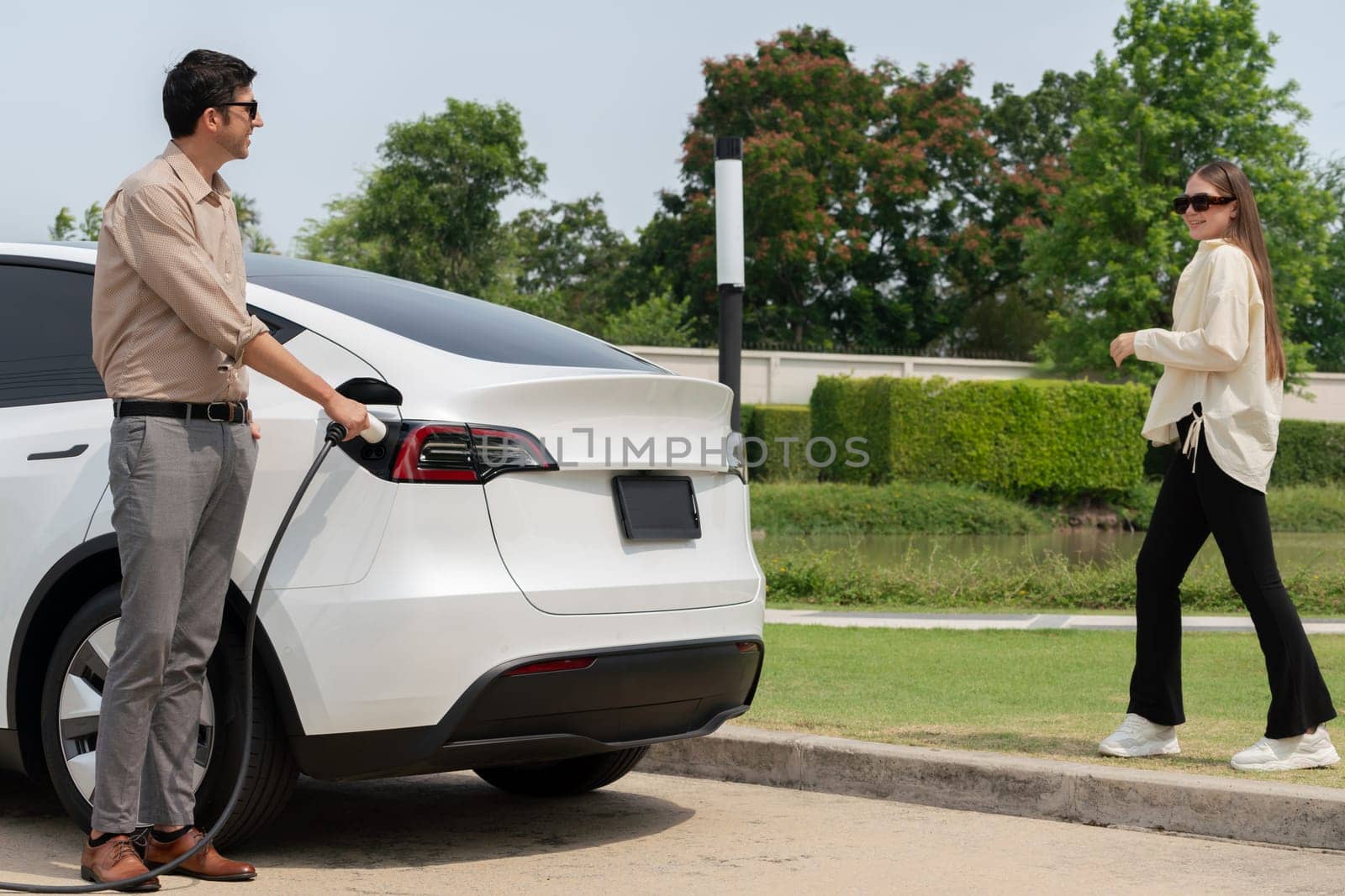 Young couple recharge electric car's battery from charging station in outdoor green city park in springtime. Rechargeable EV car for sustainable environmental friendly urban travel lifestyle.Expedient