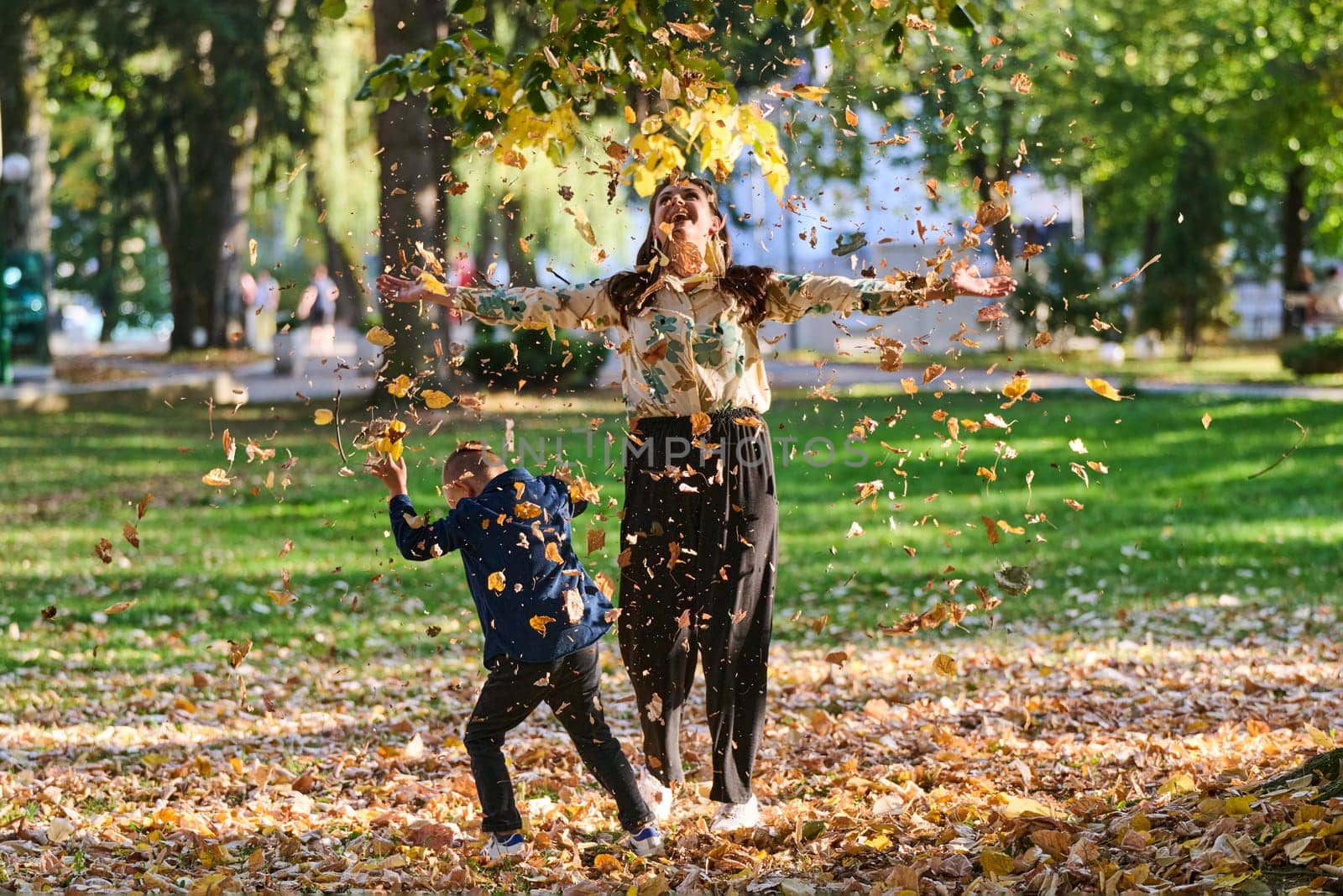 A modern woman joyfully plays with her son in the park, tossing leaves on a beautiful autumn day, capturing the essence of family life and the warmth of mother-son bonding in the midst of the fall season.