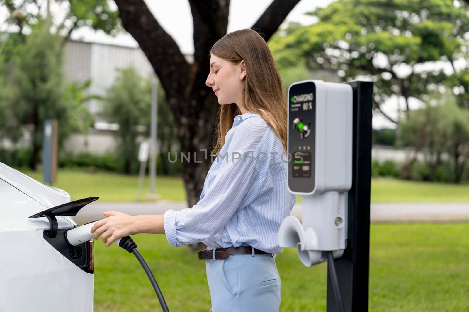 Young woman recharge EV electric vehicle's battery from EV charging station in outdoor green city park scenic. Eco friendly urban transport and commute with eco friendly EV car travel. Exalt