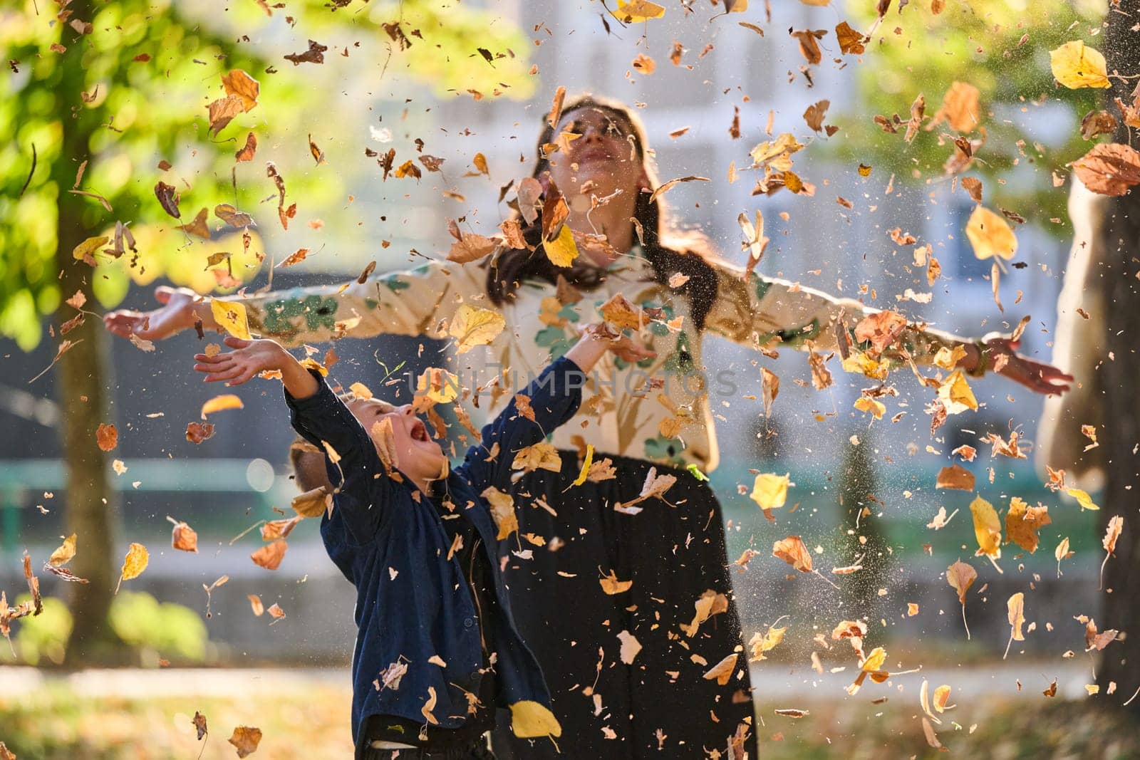 A modern woman joyfully plays with her son in the park, tossing leaves on a beautiful autumn day, capturing the essence of family life and the warmth of mother-son bonding in the midst of the fall season by dotshock