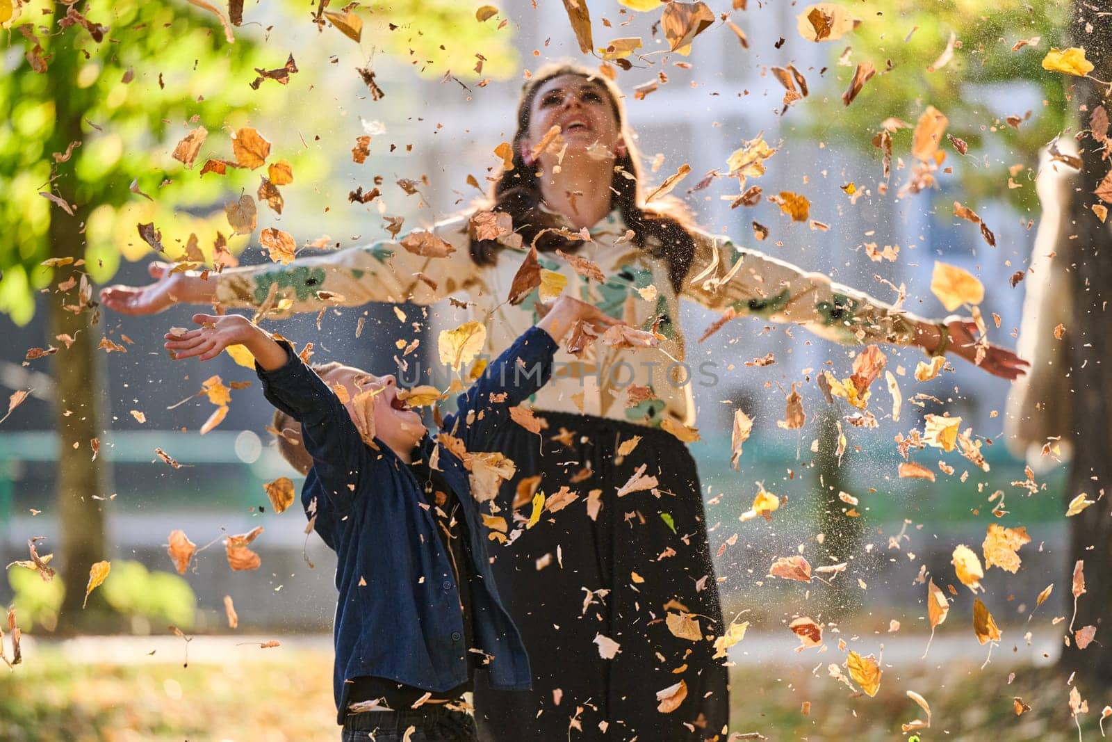 A modern woman joyfully plays with her son in the park, tossing leaves on a beautiful autumn day, capturing the essence of family life and the warmth of mother-son bonding in the midst of the fall season.