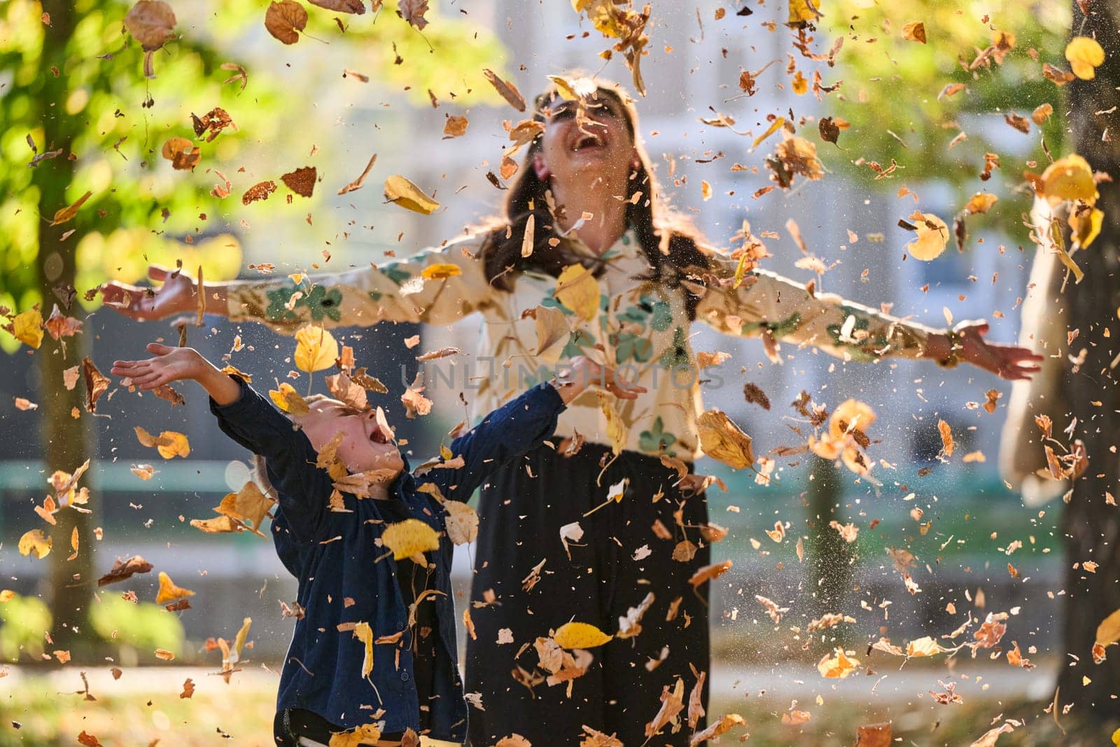 A modern woman joyfully plays with her son in the park, tossing leaves on a beautiful autumn day, capturing the essence of family life and the warmth of mother-son bonding in the midst of the fall season.