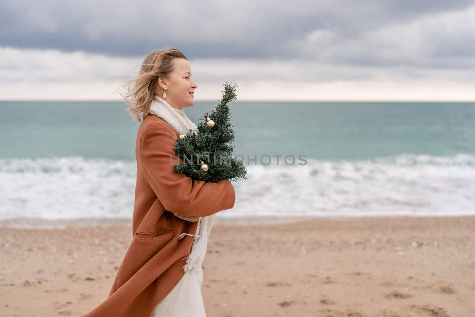 Blond woman Christmas tree sea. Christmas portrait of a happy woman walking along the beach and holding a Christmas tree on her shoulder. She is wearing a brown coat and a white suit. by Matiunina