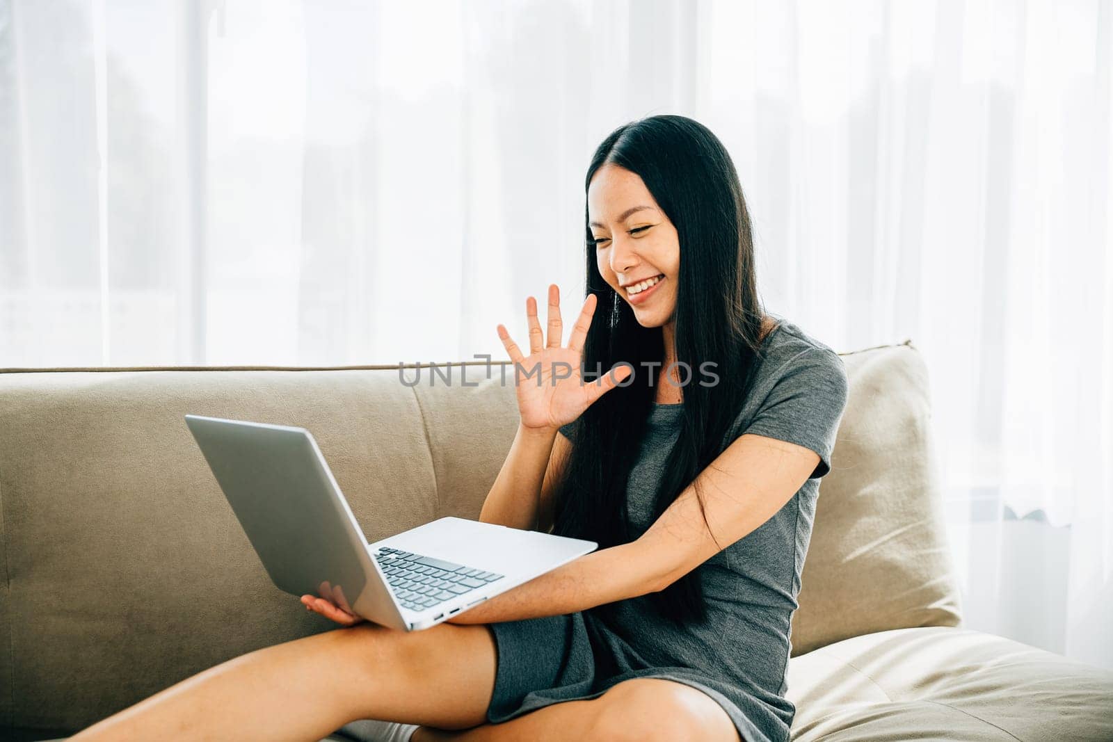 Young woman on sofa using laptop waves during video call. Millennial teacher smiles saying hello greeting clients. Engaging in distant meeting enjoying communication.