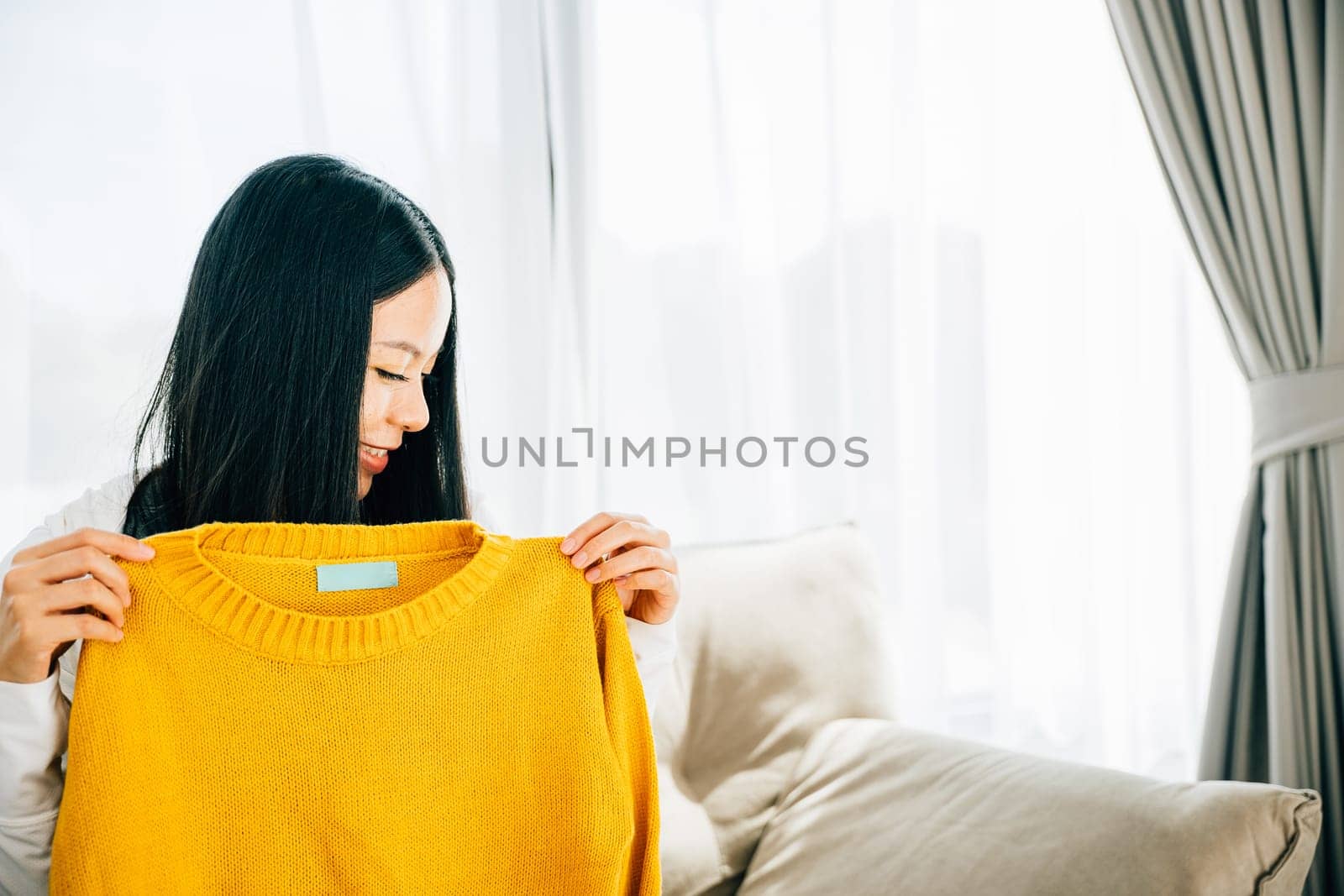 Happy Asian woman at home opens a box with a shirt smiling excitedly. Satisfied shopper unboxes online order displaying new clothes. Delivery and shopping from home concept.