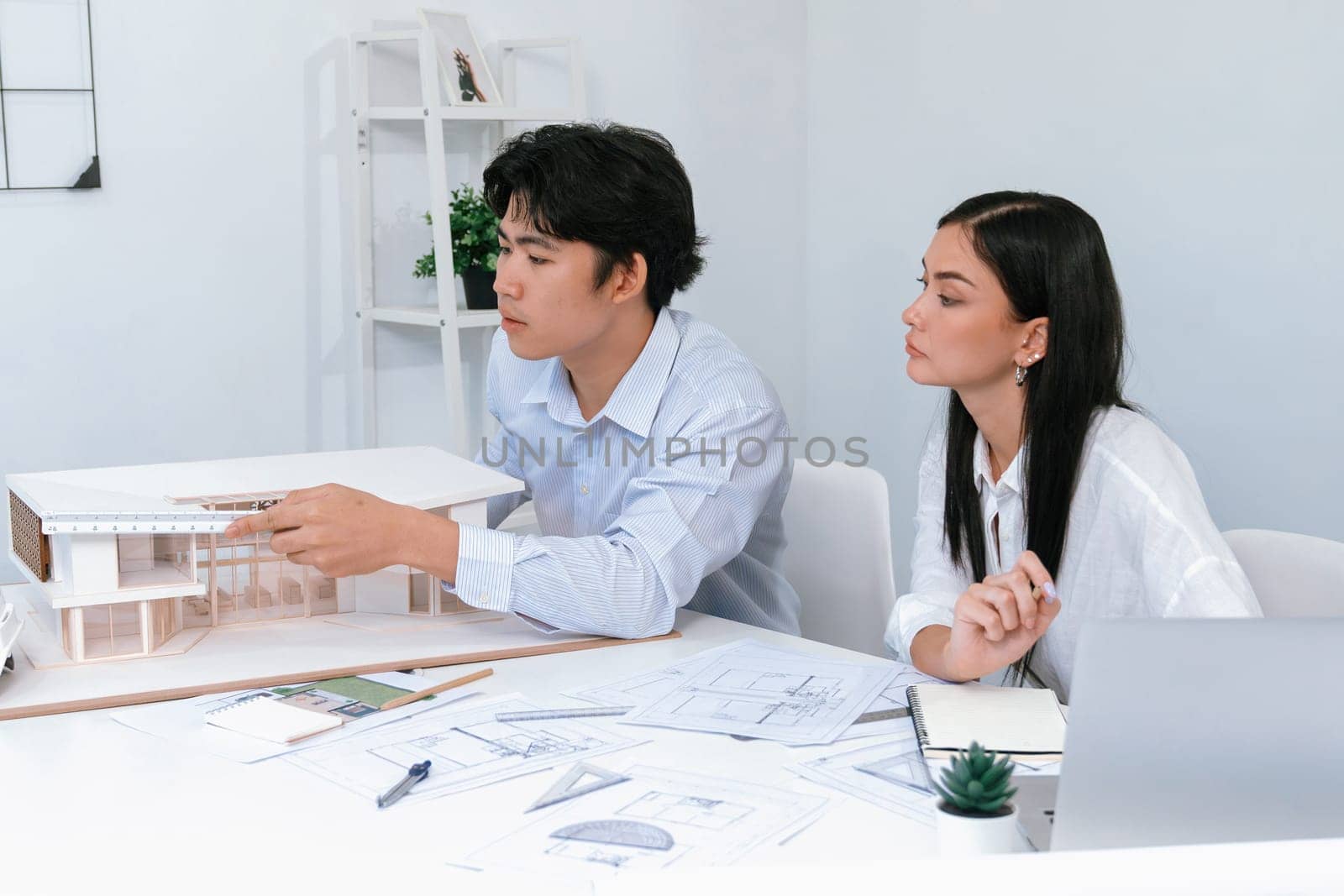 Professional male asian architect using ruler to measure house model length while young beautiful caucasian colleague using laptop to analyzed data on meeting table with house model. Immaculate.