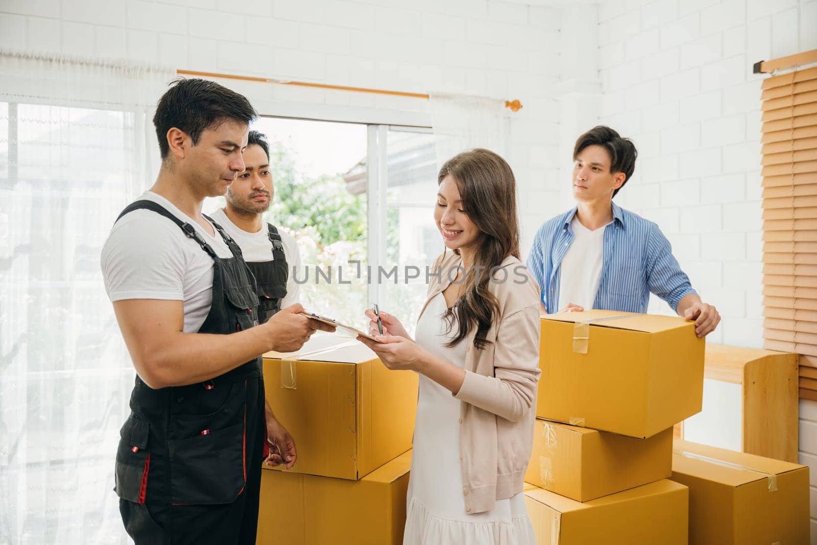 Couple inspects checklist before signing moving papers. Worker carries a box. Professional movers ensure seamless home relocation. Teamwork and efficiency displayed. Movind Day to New home