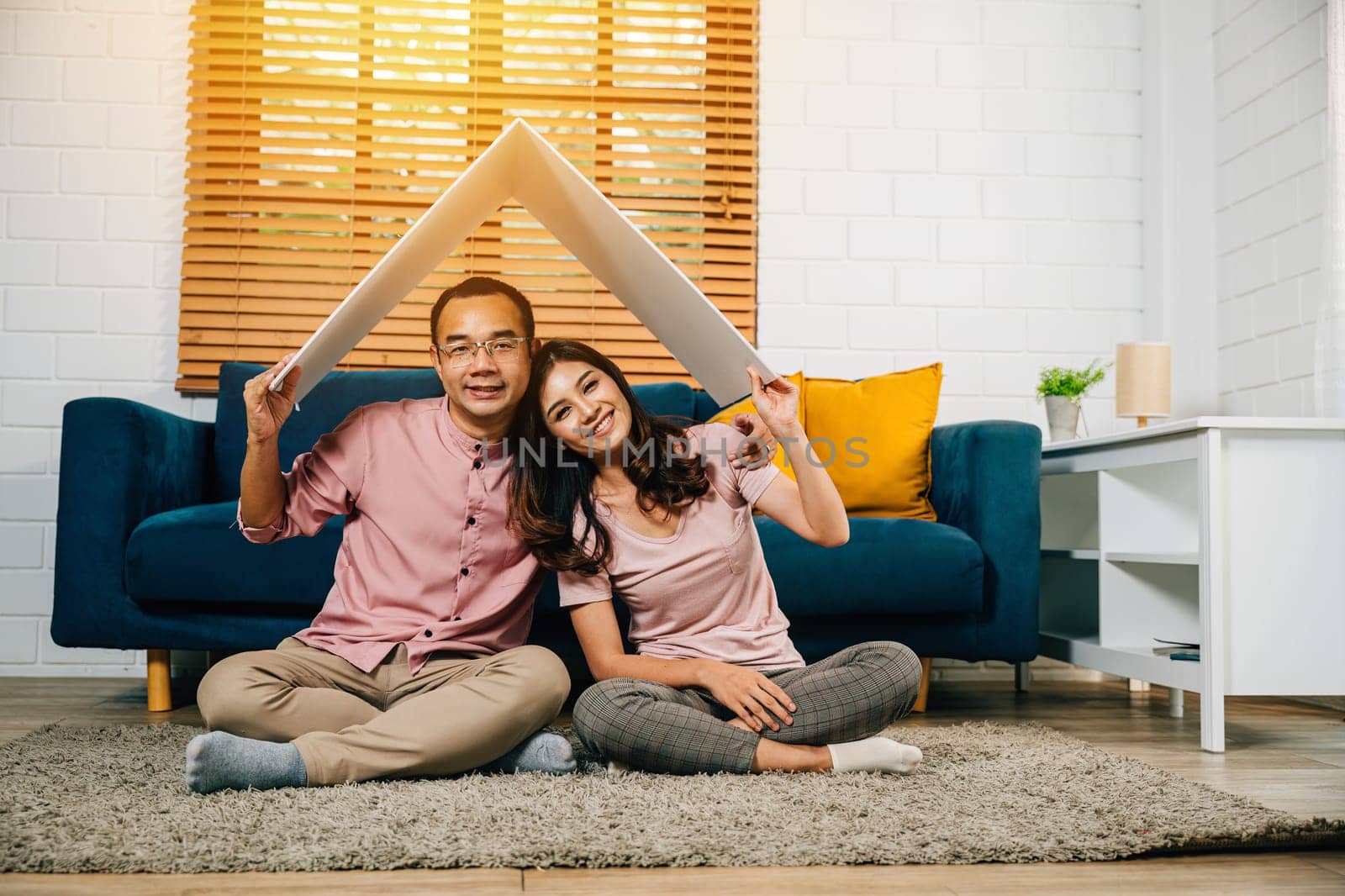 In their new empty apartment Asian husband and wife create cardboard roof celebrating their achievement and happiness. Full-length portrait against white studio wall showing their security and bonding