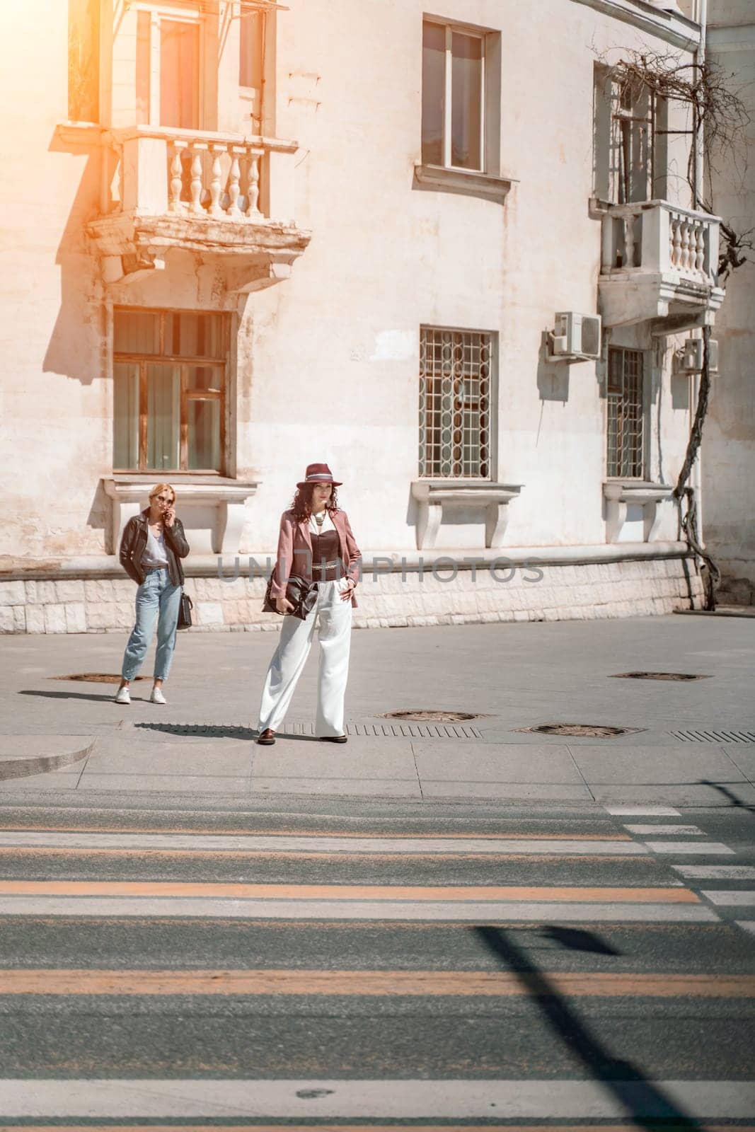 Woman city road crossing. Stylish woman in a hat crosses the road at a pedestrian crossing in the city. Dressed in white trousers and a jacket with a bag in her hands