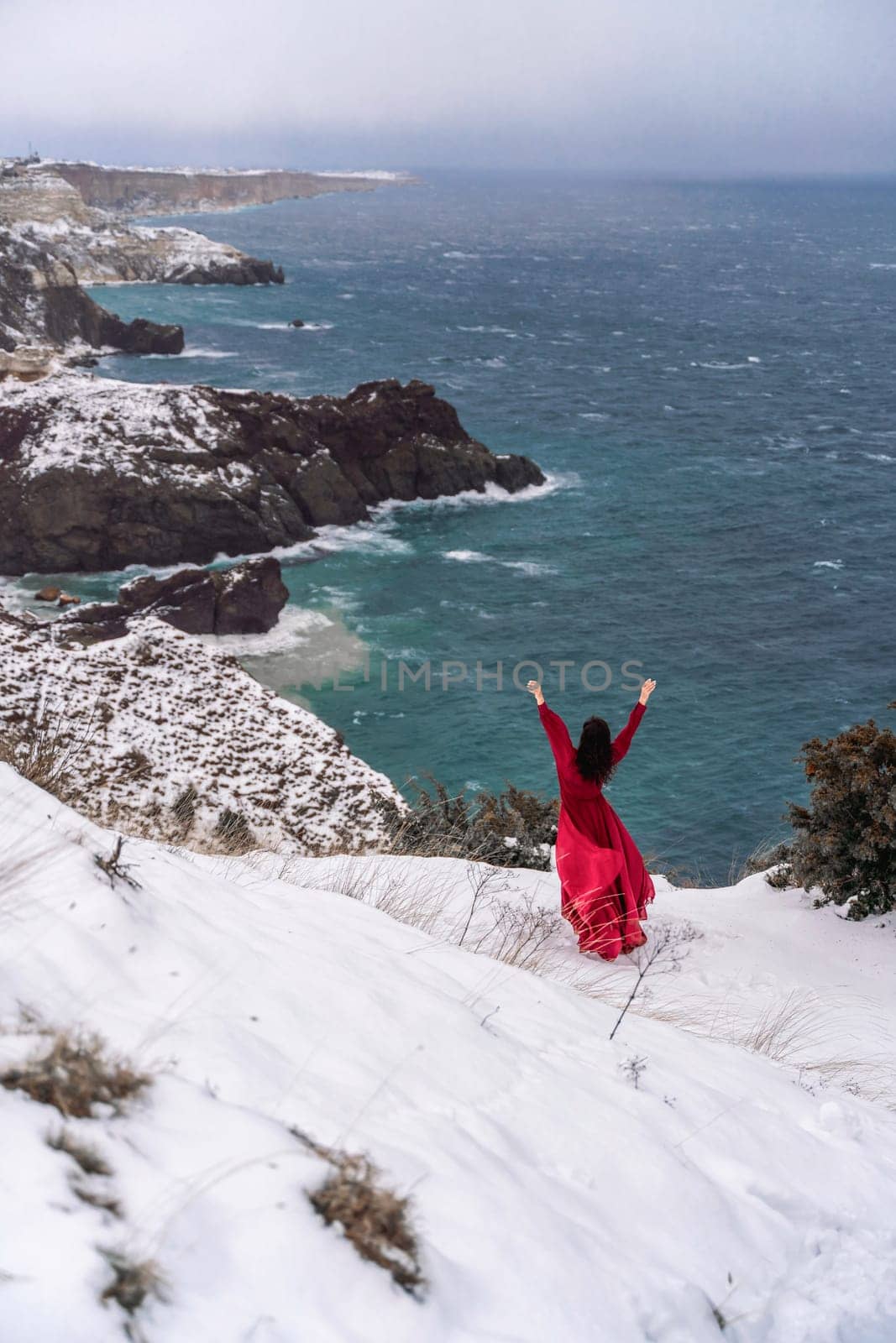 Woman red dress snow sea. Happy woman in a red dress in the snowy mountains by the emerald sea. The wind blows her clothes, posing against sea and snow background. by Matiunina