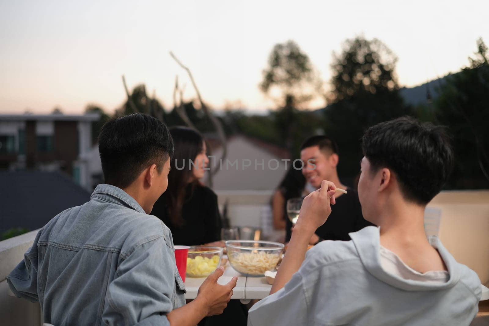 Rear view of young people enjoying conversation at rooftop party in evening.