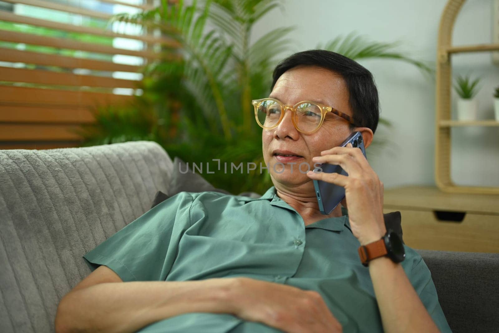 Carefree middle age man in glasses having pleasant phone conversation while relaxing on couch at home by prathanchorruangsak