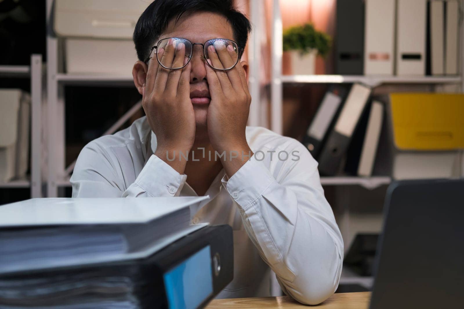 Exhausted young man with laptop in office working late sitting on desk in office overtime at night. Frustrated young businessman massaging his nose and keeping eyes closed while sitting at his working place in office.