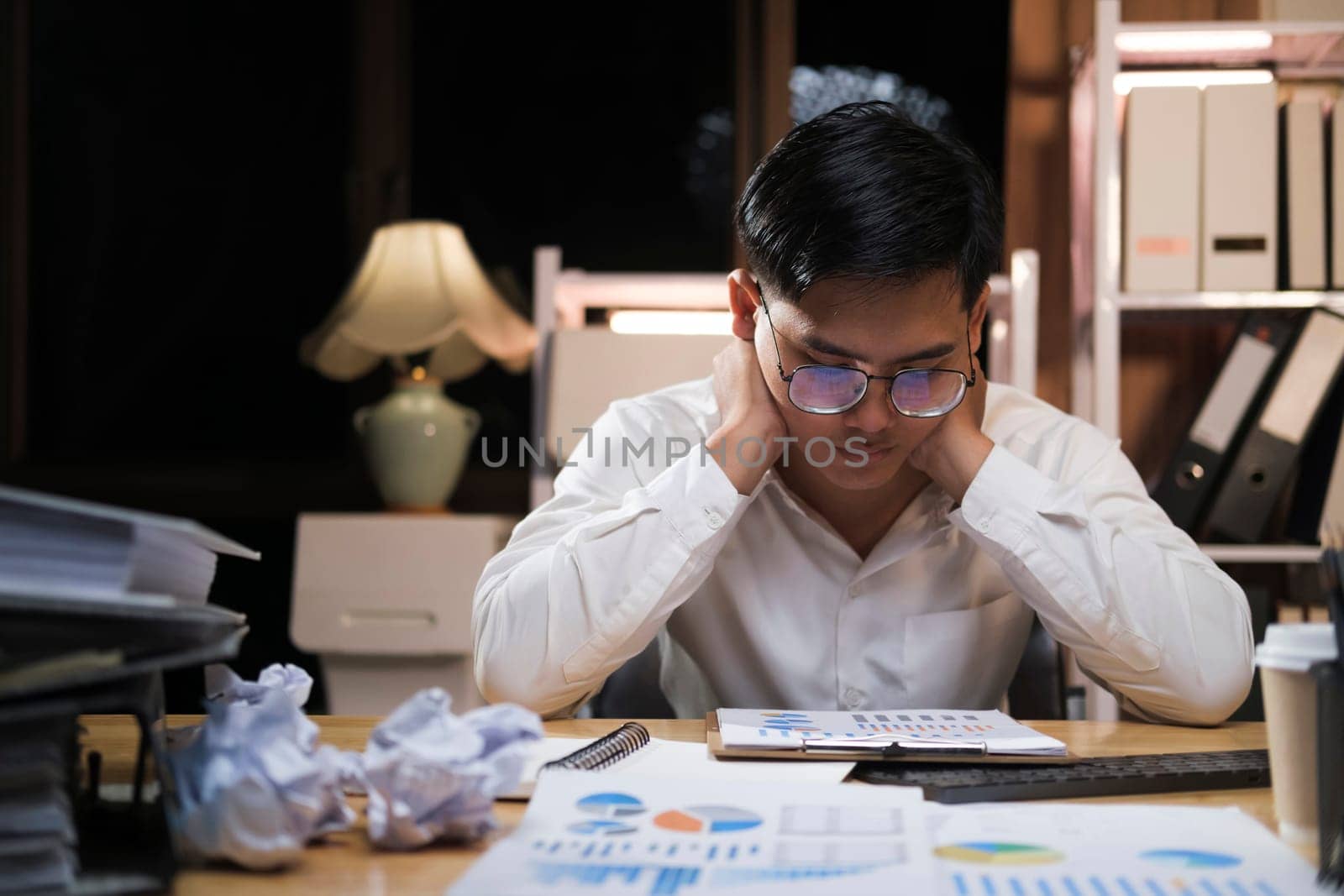 Asian man working late sitting on desk in office overtime at night. Business man feeling tired and stress for overload job.