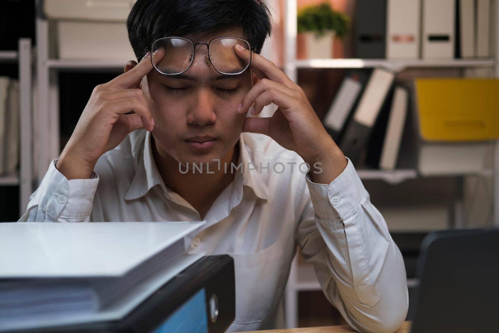 Exhausted young man with laptop in office working late sitting on desk in office overtime at night. Frustrated young businessman massaging his nose and keeping eyes closed while sitting at his working place in office.