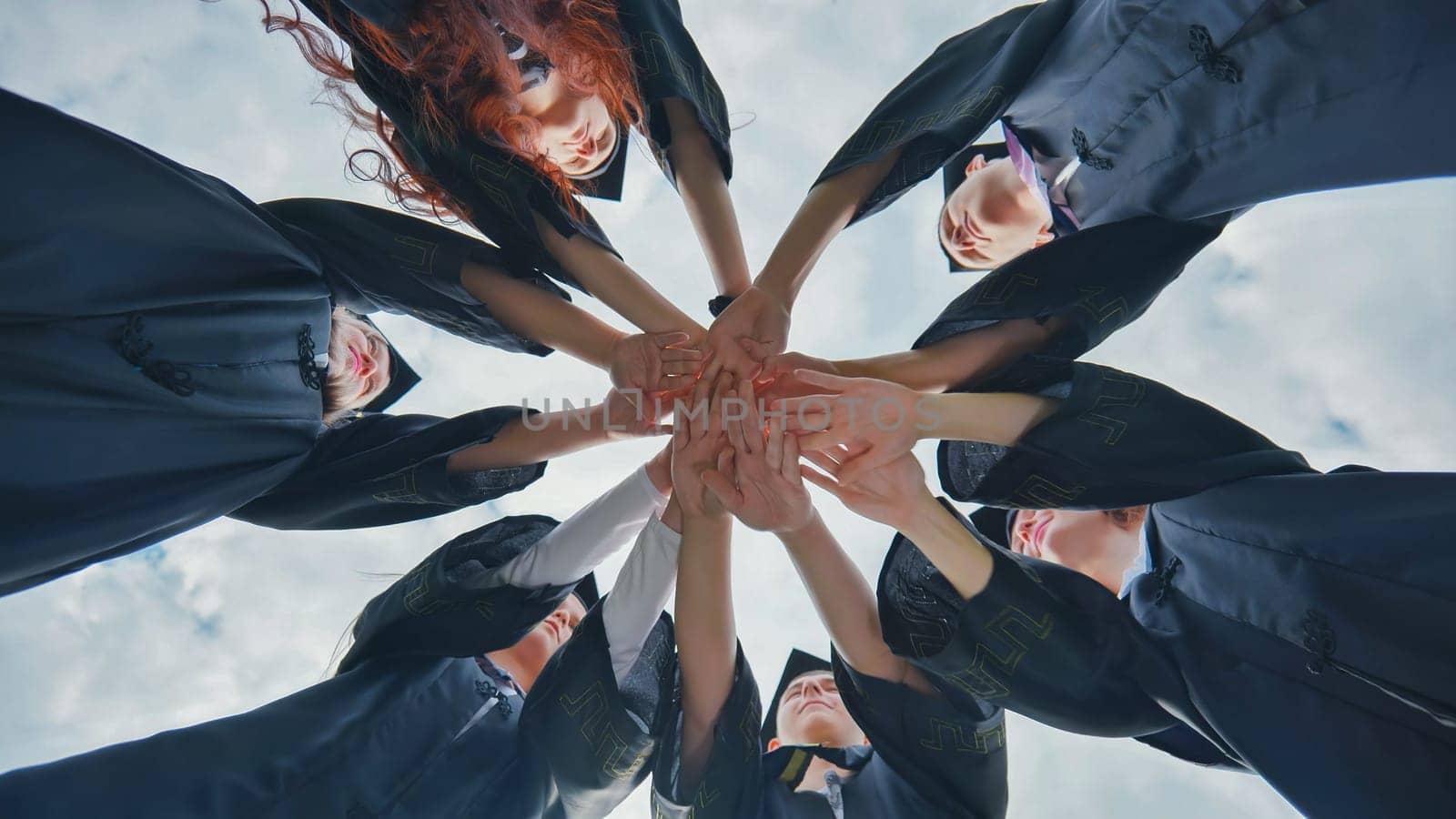 Team of college or university students celebrating graduation. Group of happy successful graduates in academic hats and robes standing in circle and putting their hands together. by DovidPro