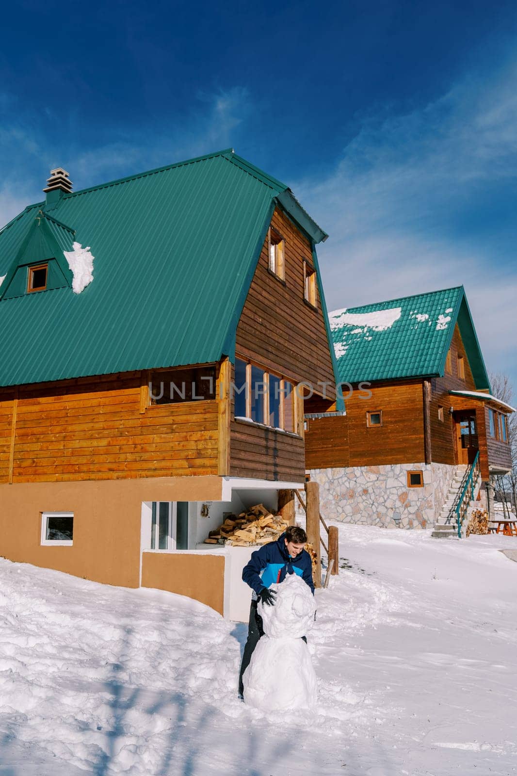 Young man makes a snowman in the yard next to the wooden cottages. High quality photo