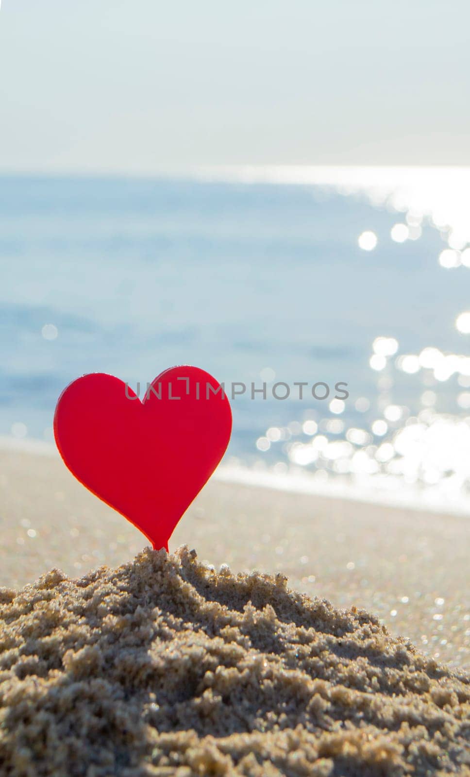 Plastic stick in shape of red heart in sand on sandy beach of sea shore on background of sea waves on sunny summer day close-up. Concept Love relationship romance amorousness amour St. Valentines Day