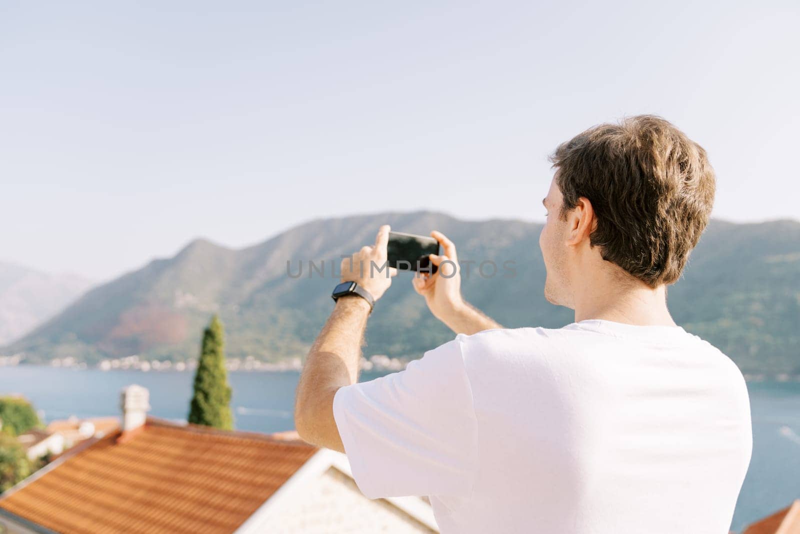 Man takes pictures of the sea above the roofs of houses with his smartphone. Back view by Nadtochiy