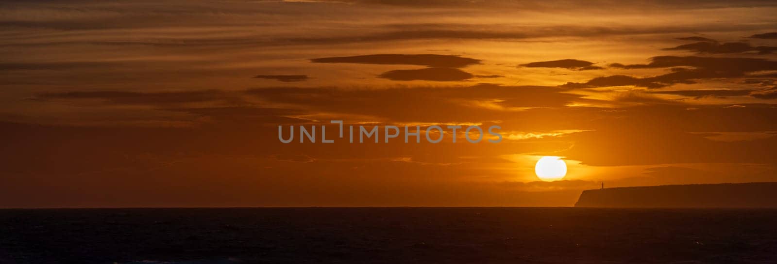 Lighthouse atop cliffs silhouetted by orange sunset over calm sea.