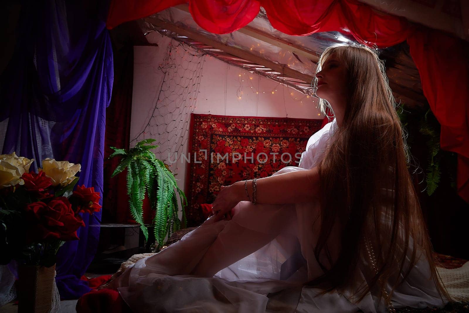 Beautiful European girl looking like Arab woman in red room with rich fabrics and carpets in sultan harem. Photo shoot of an oriental style odalisque. A model poses in sari as indian woman in india