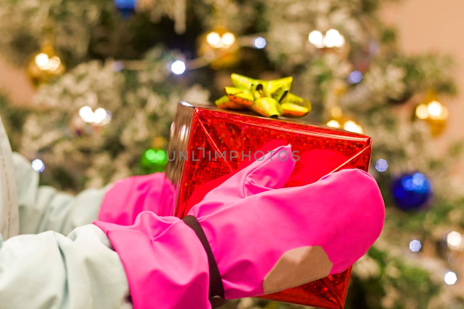 girl baby holds in pink gloves red christmas box before christmas tree.