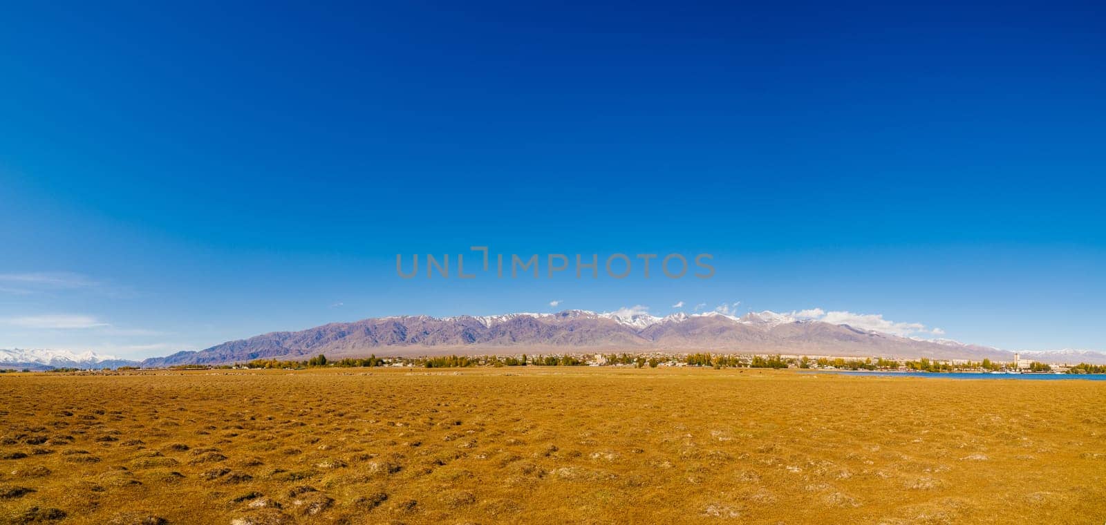 field covered with dry grass bumps with small Kyrgyz town Balykchy and high mountains on the horizon, wide angle panoramic view.