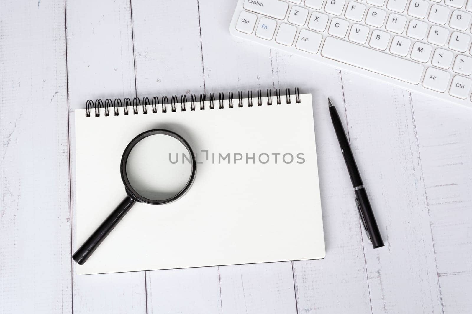 Home office workspace with white keyboard, note book, pen and magnifying glass. Flat lay, top view. Copy space.