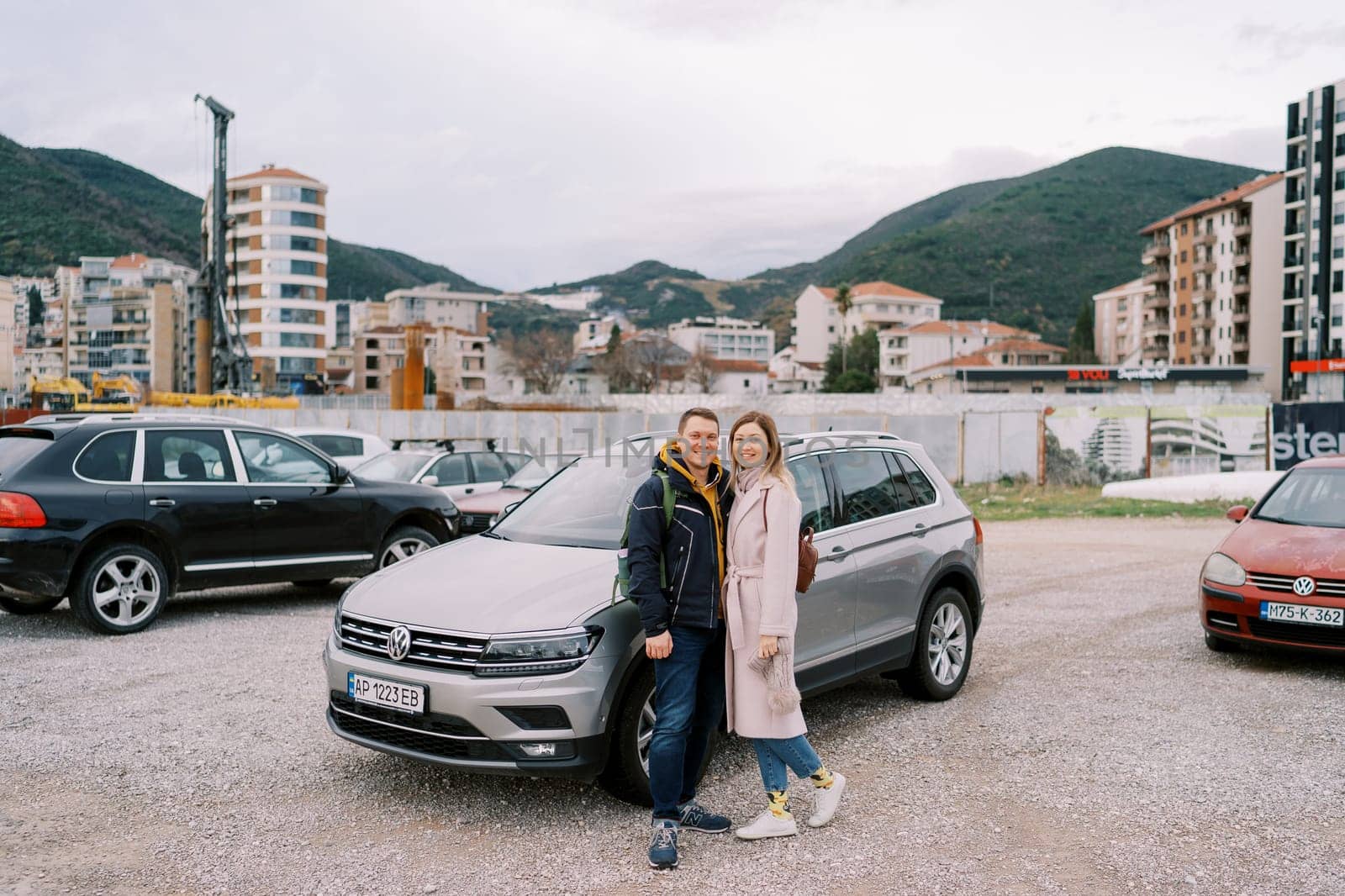 Smiling couple stands near a car in a parking lot against the backdrop of high-rise buildings at the foot of the mountains. High quality photo