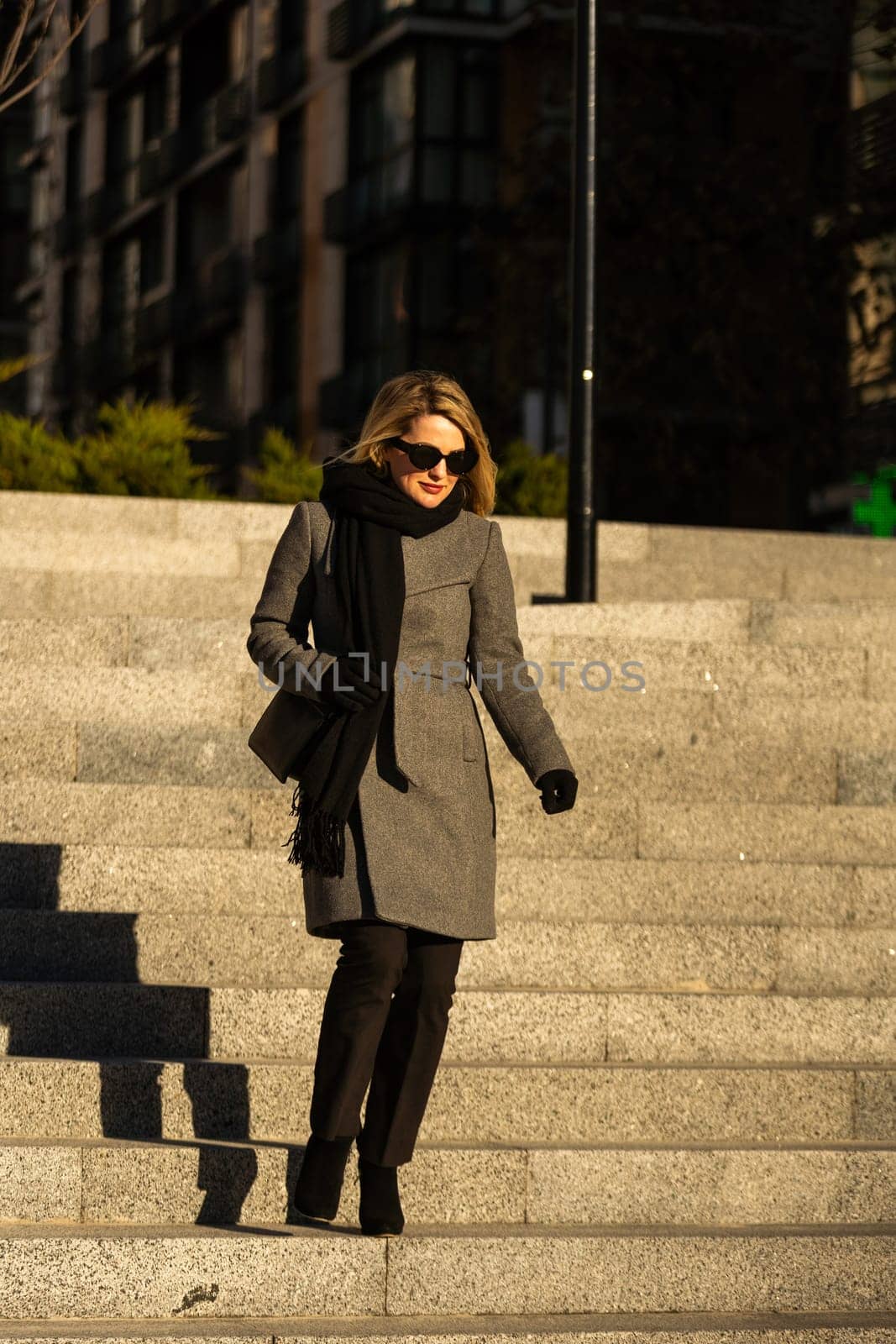 The young lady in black stylish coat with black scarf walking the sun rays at sunset on the background the cold autumn day in the city by Andelov13