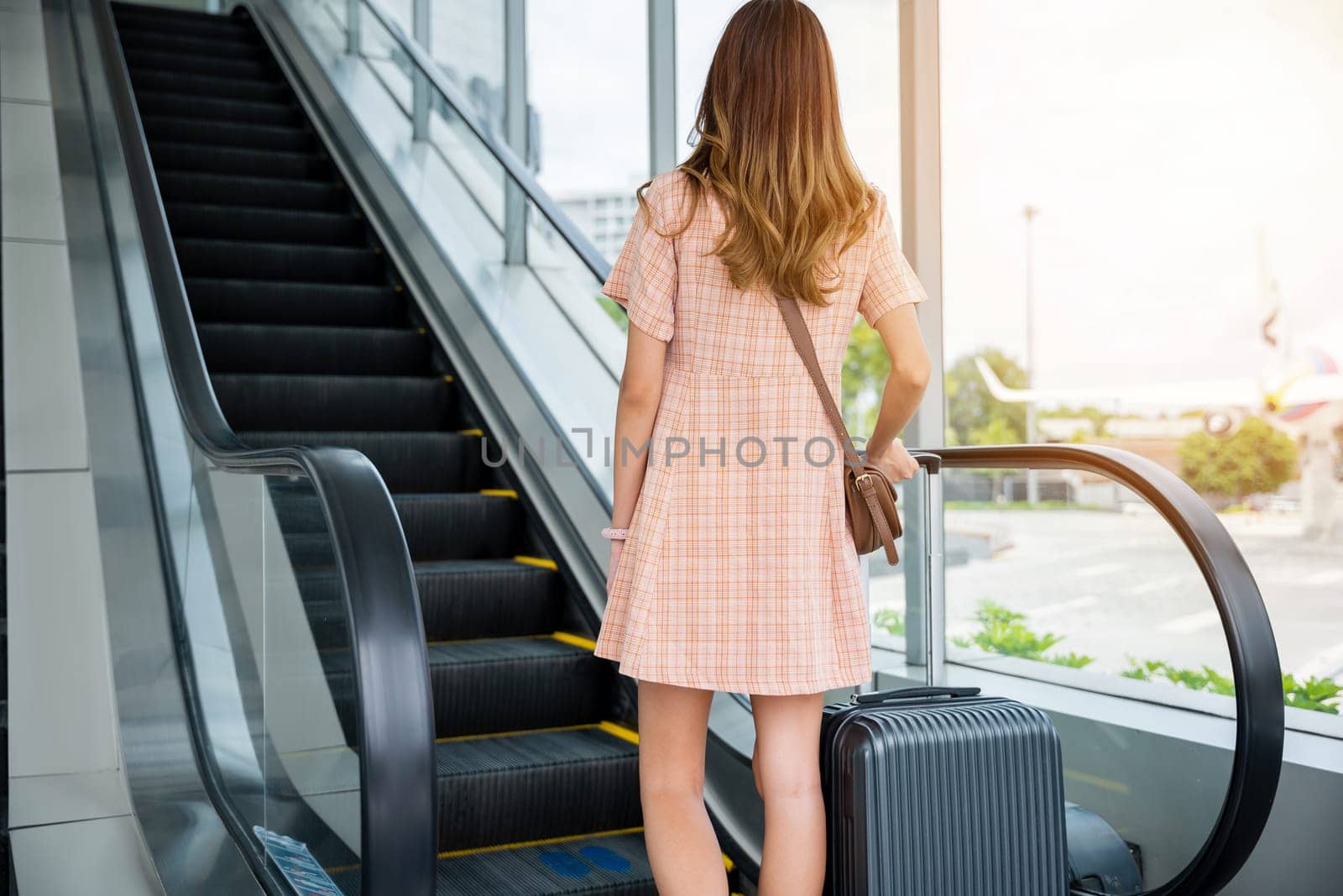 A lady in transit carries her luggage up an escalator in a bustling airport terminal. She looks forward to her destination and the exploration and attraction that awaits her