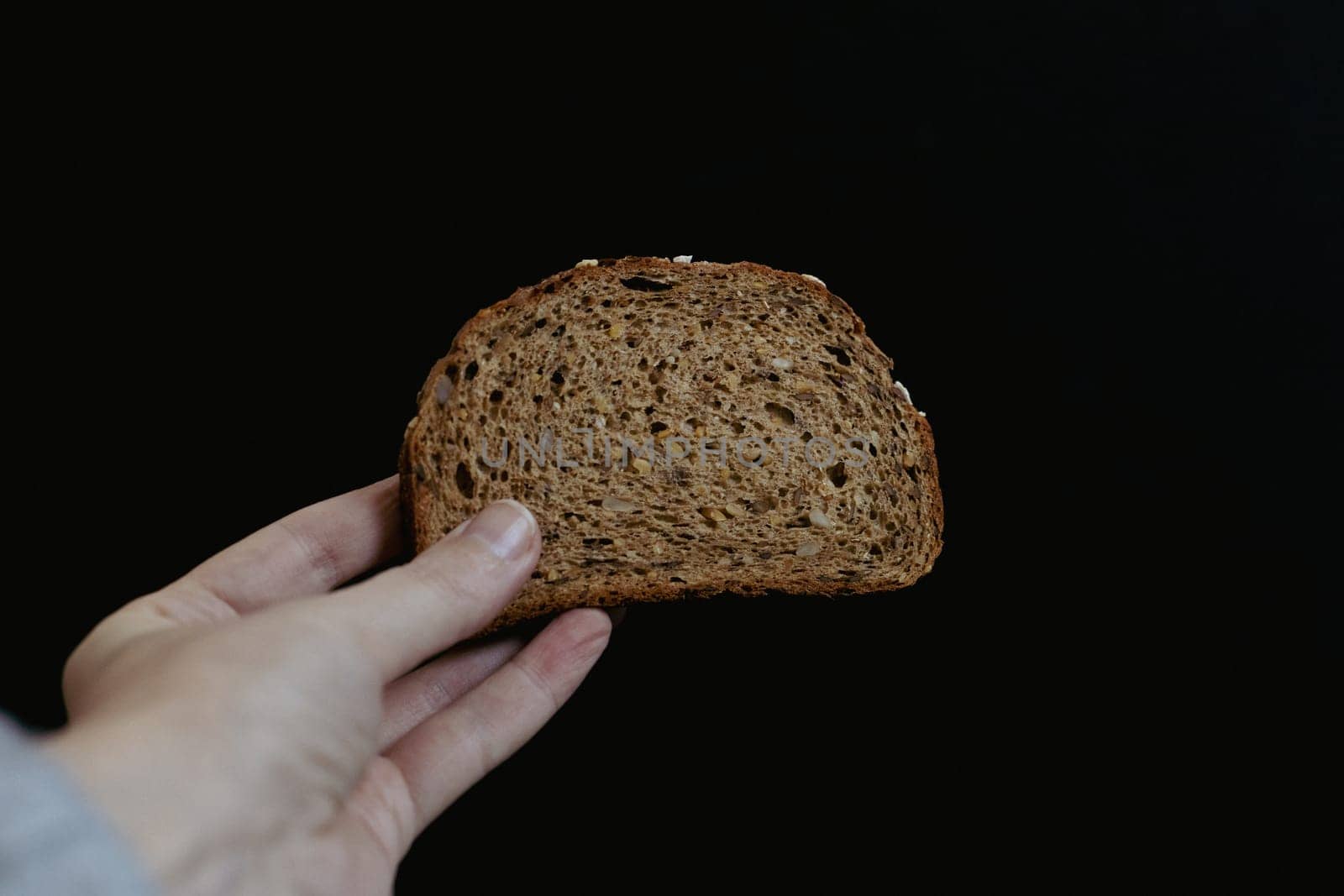 The hand of a young caucasian girl holds one piece of rye bread with sunflower seeds on a black background, close-up side view. The concept of baking bread.