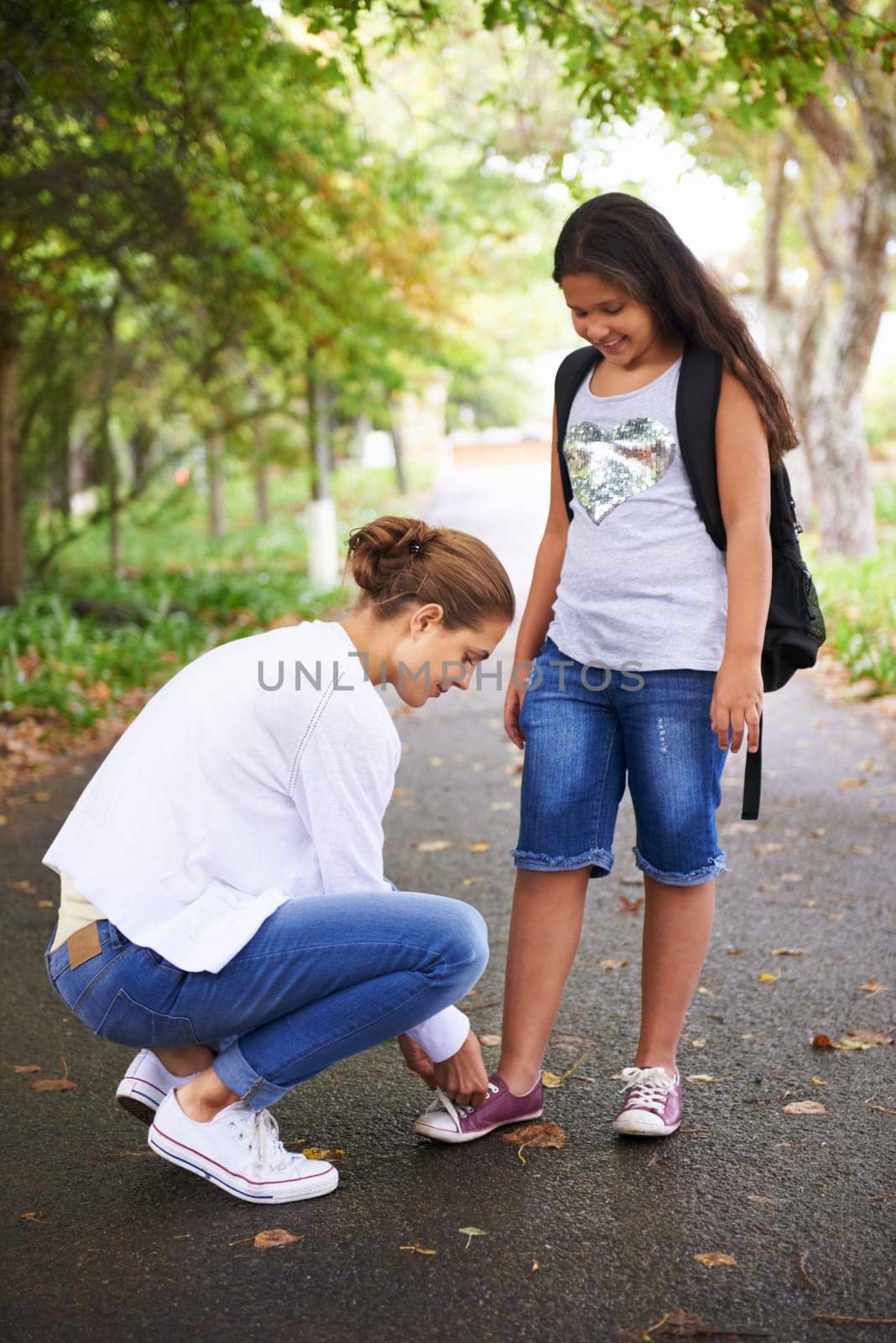 Woman, teacher and tying student shoes in park for back to school, learning or education in nature. Female person teaching little girl to tie laces on asphalt path, road or outdoor street in woods.