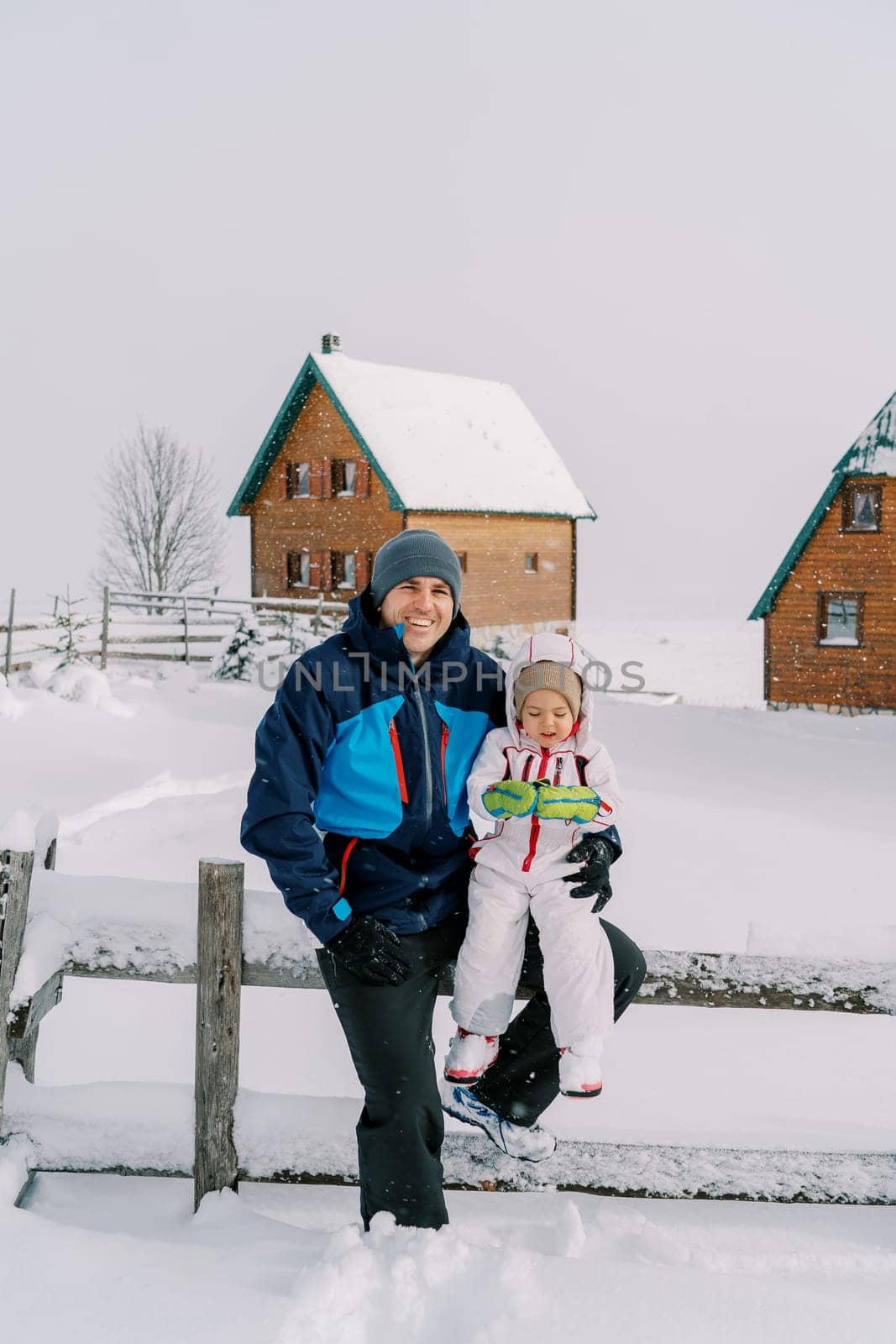 Smiling dad with a little girl on his lap sitting on a snow-covered fence in the village. High quality photo