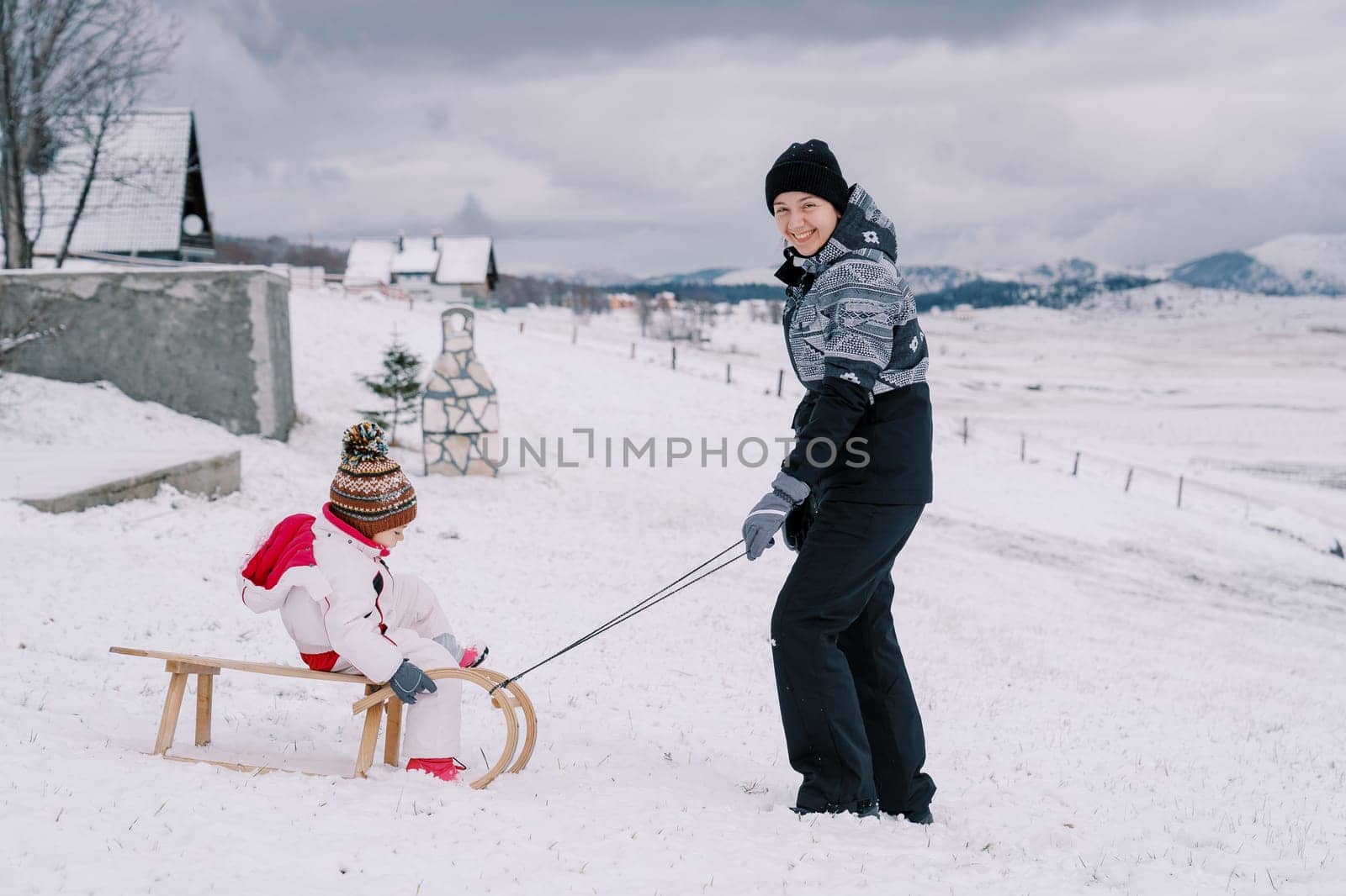 Smiling mother pushing a wooden sled with a little girl down a snowy hill. High quality photo
