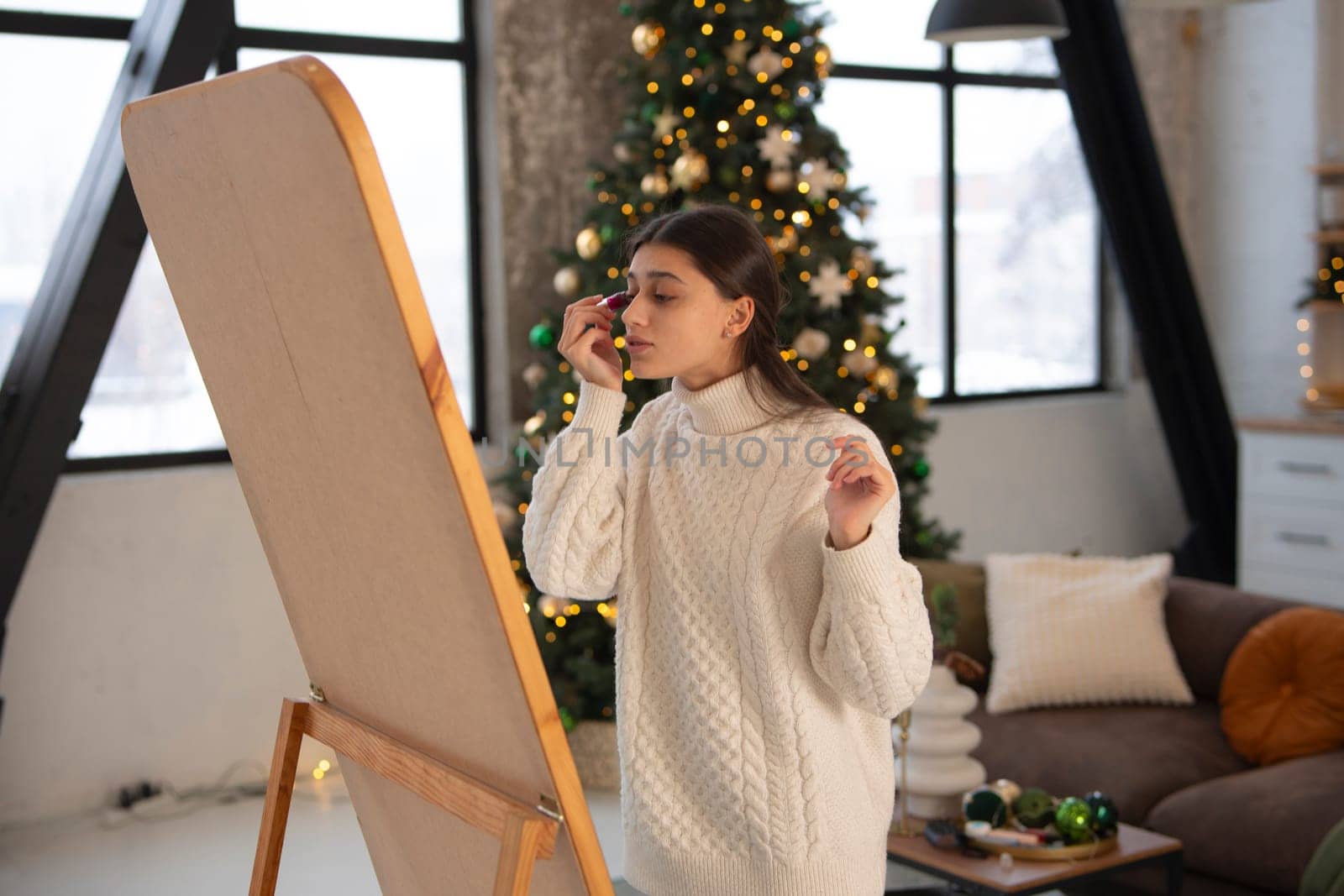 Creating a festive mood, a beautiful young woman does makeup in a Christmas setting. by teksomolika