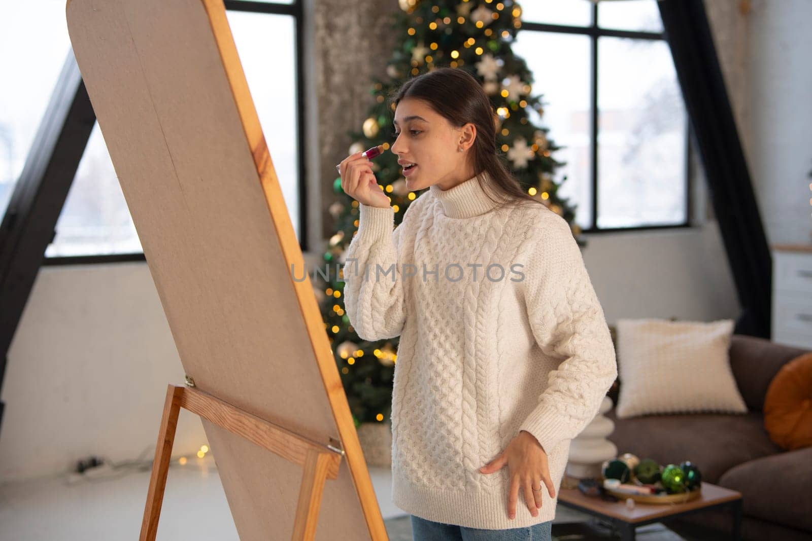 In a Christmas ambiance, a lovely young lady applies makeup before the mirror. High quality photo